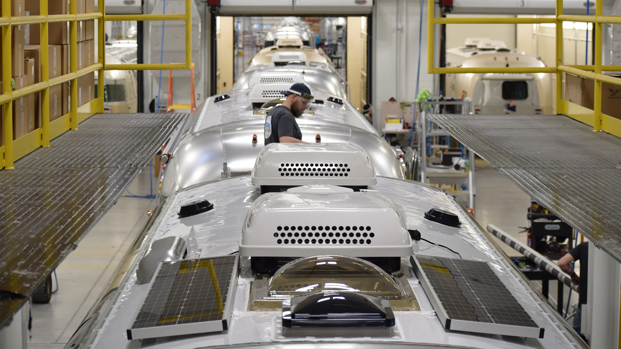 A worker inspects the roof of a silver travel trailer in a factory, surrounded by several other similar trailers lined up in a row inside the production facility.