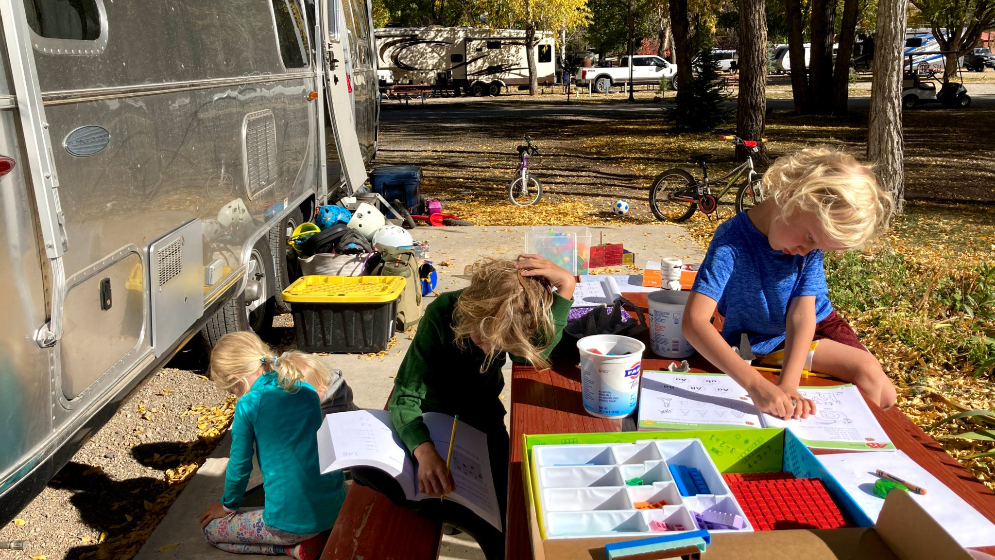 Three young children sit at a picnic table next to an RV, coloring and doing activities with books and supplies. Outdoor items, bikes, and trees are visible in the background on a sunny day.