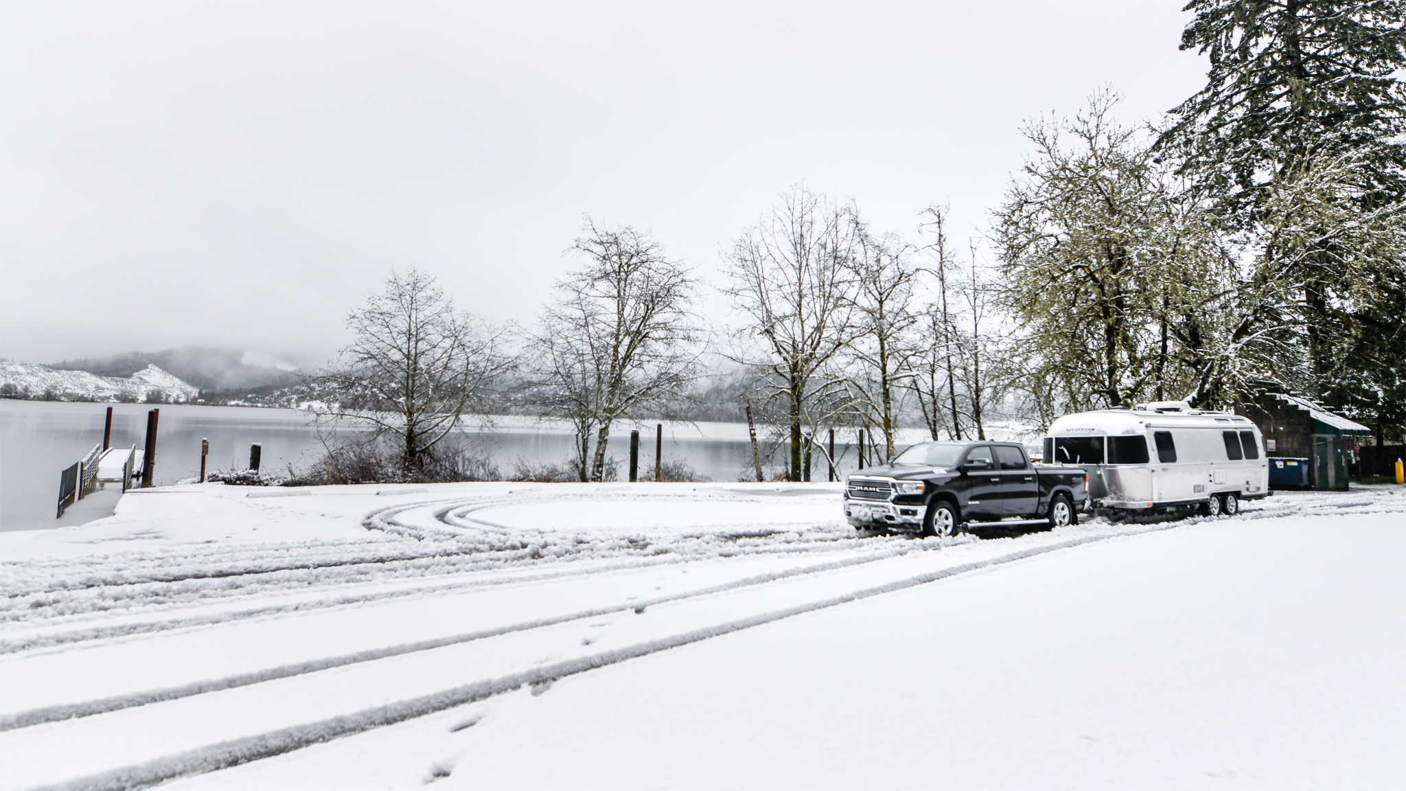 A silver truck towing an Airstream trailer is parked on a snowy lakeside road, with snow-covered trees and tire tracks visible, and hills in the background under an overcast sky.