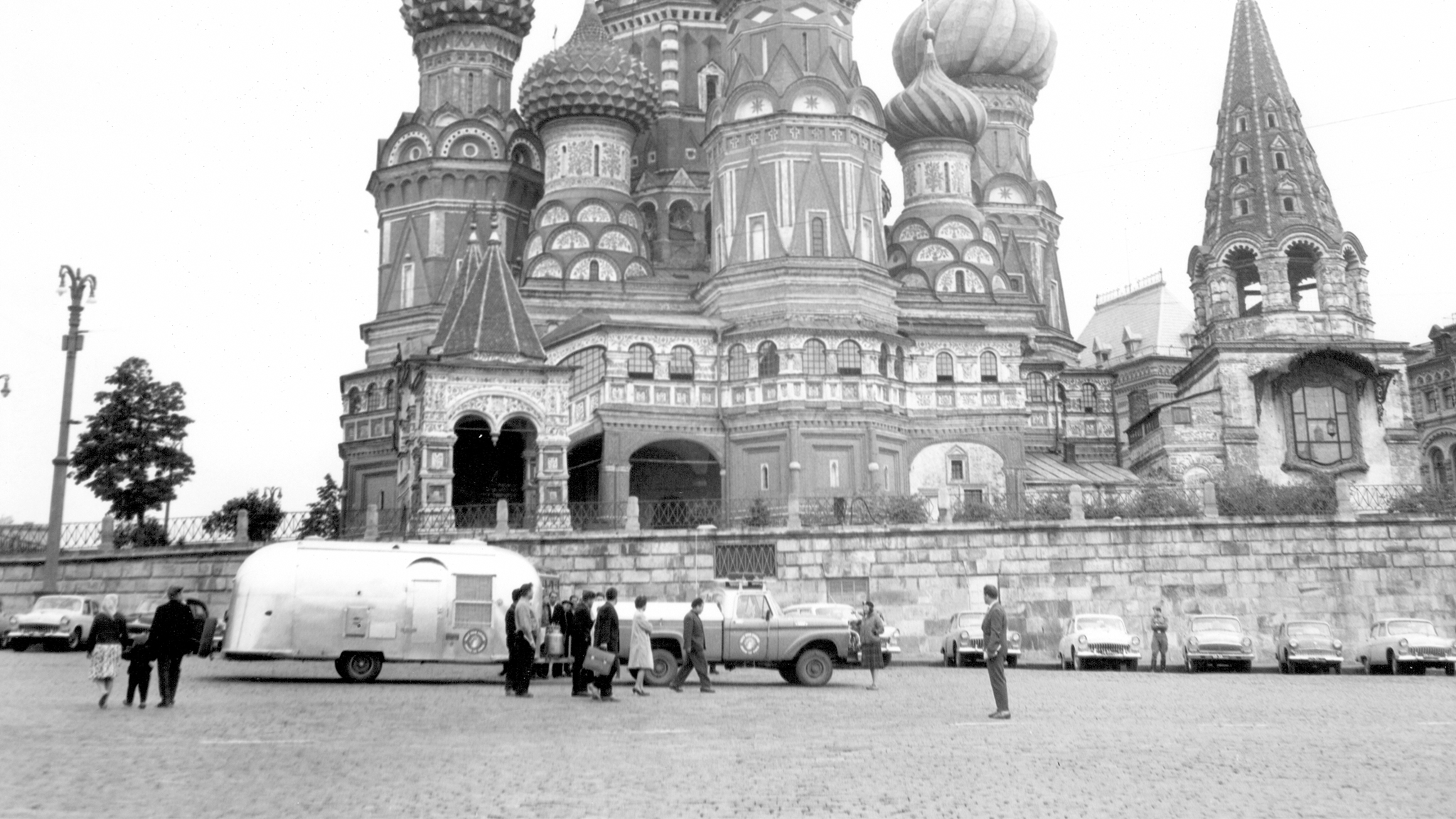 Black and white photo of St. Basil's Cathedral in Moscow, with people gathered near a trailer and vintage cars parked nearby.