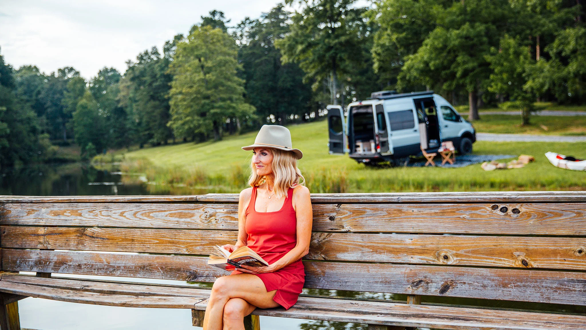 Woman in a red dress and hat reading a book on a wooden bench by a lake, with a camper van and trees in the background.
