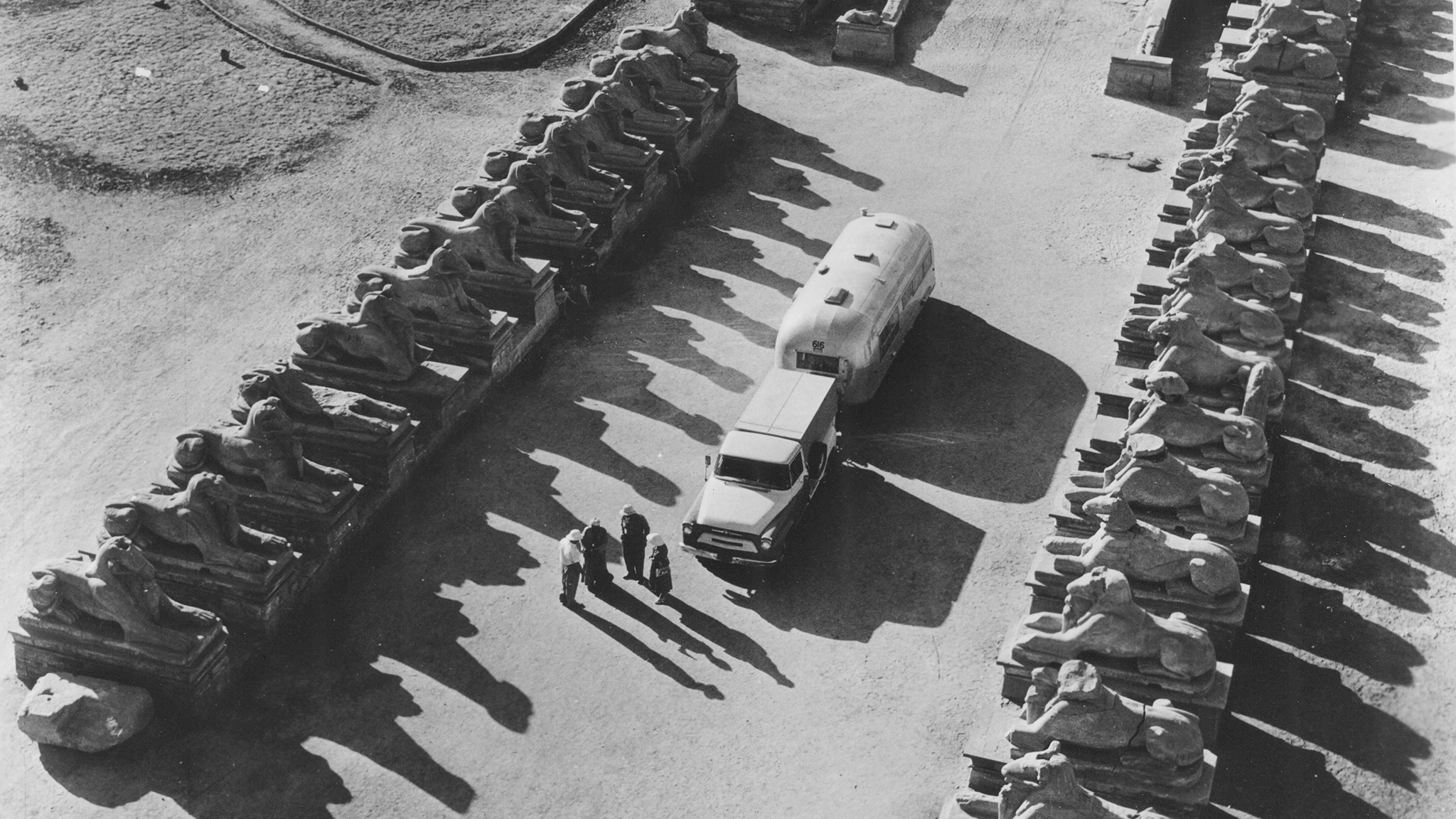 Aerial view of a vintage car and trailer driving through rows of stone sphinx statues, with four people walking nearby, casting long shadows.