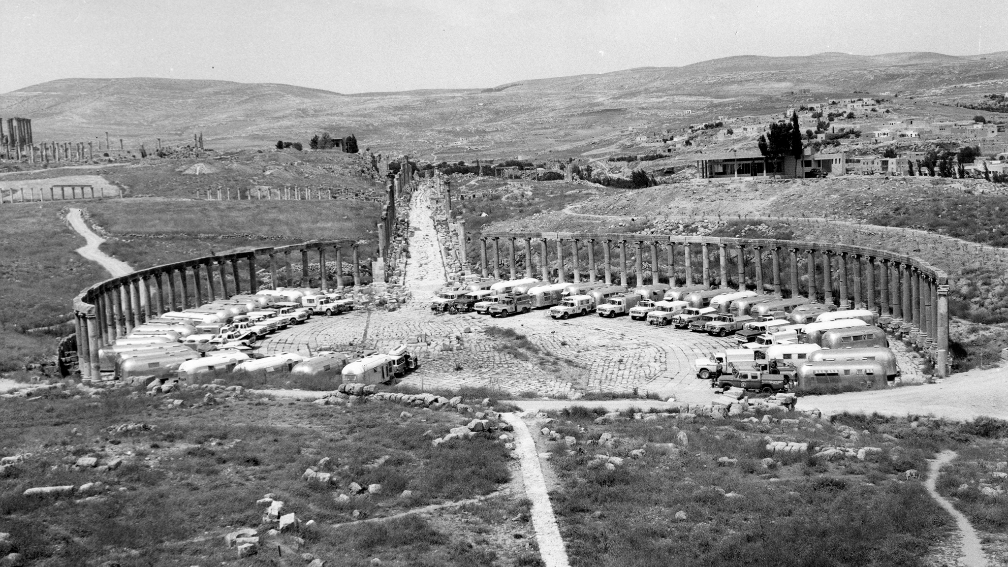 Black and white aerial view of the oval forum in the ancient city of Jerash, Jordan, with many trucks and vehicles parked inside the colonnaded structure; hills and scattered buildings in the background.