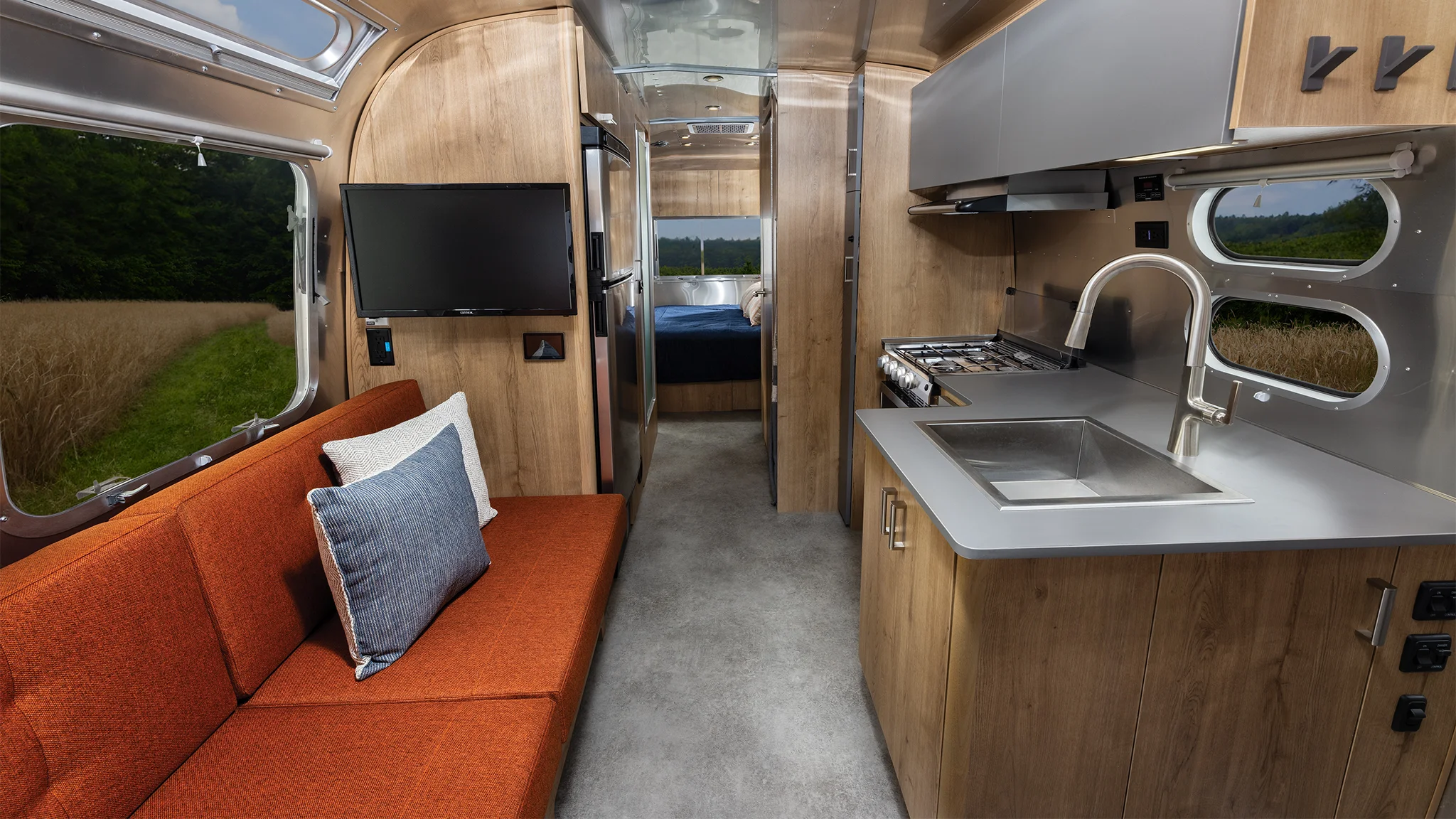 Interior of a modern RV with a red sofa, TV, kitchen area with sink and stove, and a bed in the background. Large windows show outdoor scenery.