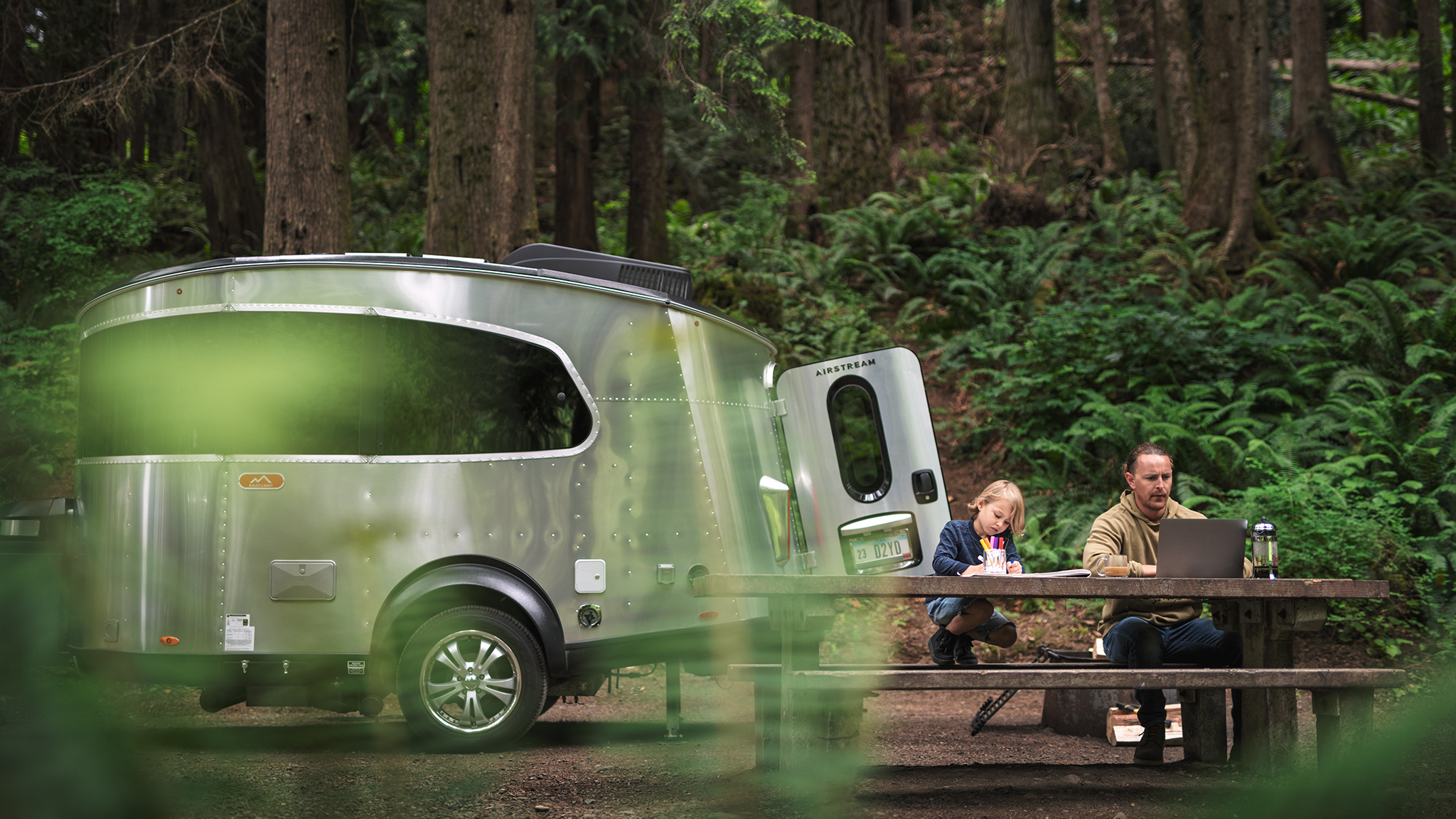 A man works on a laptop and a child draws at a wooden picnic table beside a silver camper trailer in a forested campsite with tall trees and green ferns.