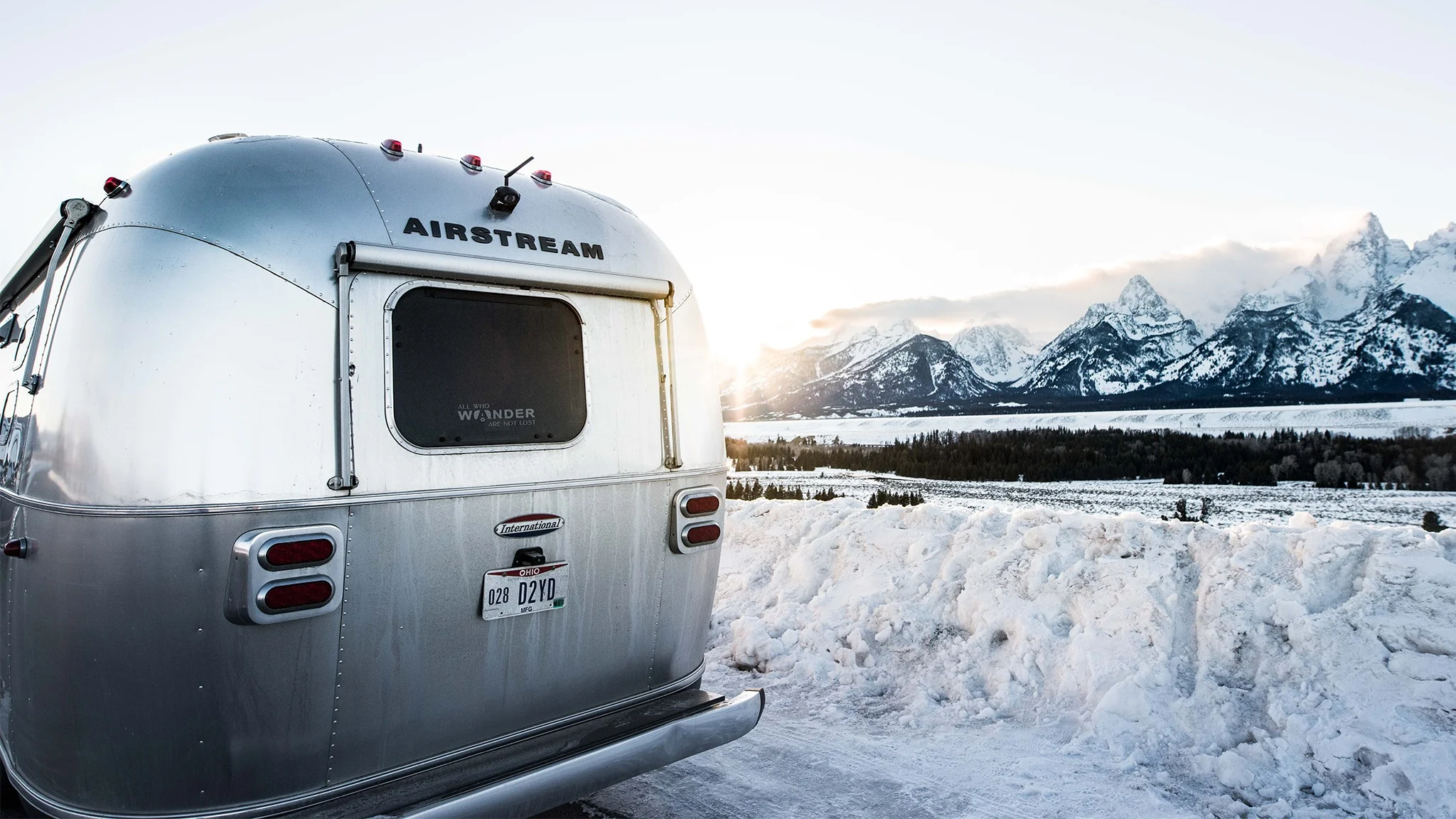 A silver Airstream trailer parked on a snowy roadside overlooks a mountain range, with the sun setting in the background.