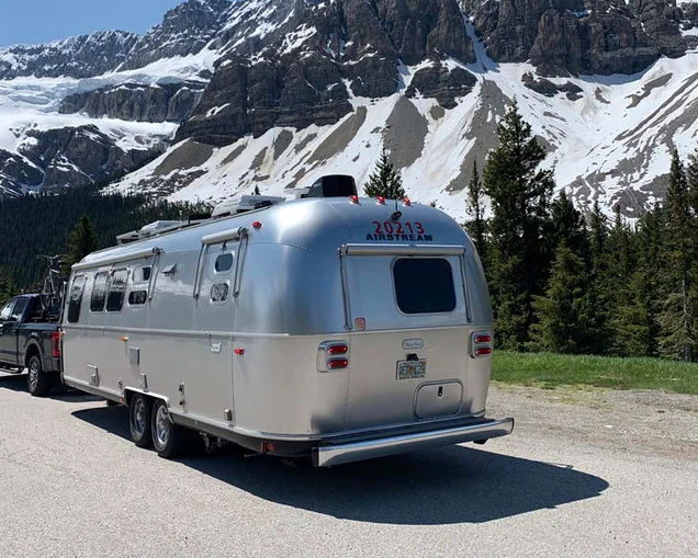 Airstream camper being towed by a truck through snowy mountains.|Airstream camper being towed by a truck through snowy mountains.|Airstream Flying Cloud Office camper boondocking.|A man sitting in the Airstream Flying Cloud Office writing music.|A space in the Flying Cloud Office camper made as a spot for a keyboard.|A man working from anywhere in his Airstream Flying Cloud office camper.|Scott and Kim posing for an image next to a lake and mountains.|Airstream Flying Cloud camper parked at a campground with trees surrounding the trailer.