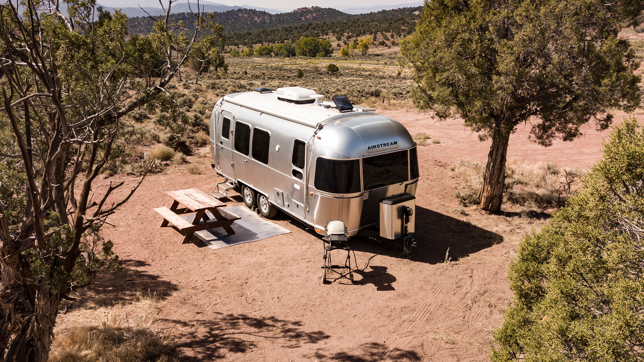 A silver Airstream trailer is parked on dry, sandy ground surrounded by trees in a rural, desert-like area. Nearby, there is a wooden picnic table and bench, with mountains visible in the background.