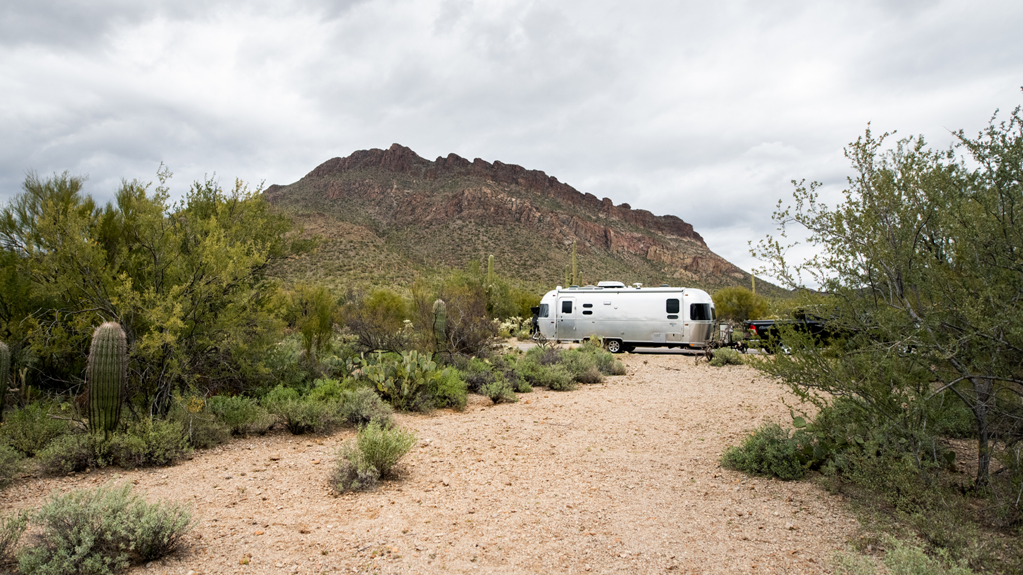An Airstream boondocking in the dessert and is hooked up to a truck.|An Airstream boondocking in the dessert and is hooked up to a truck.|A woman carrying a cup to sit down by the campfire that is made while camping in her Airstream travel trailer in the desert.
