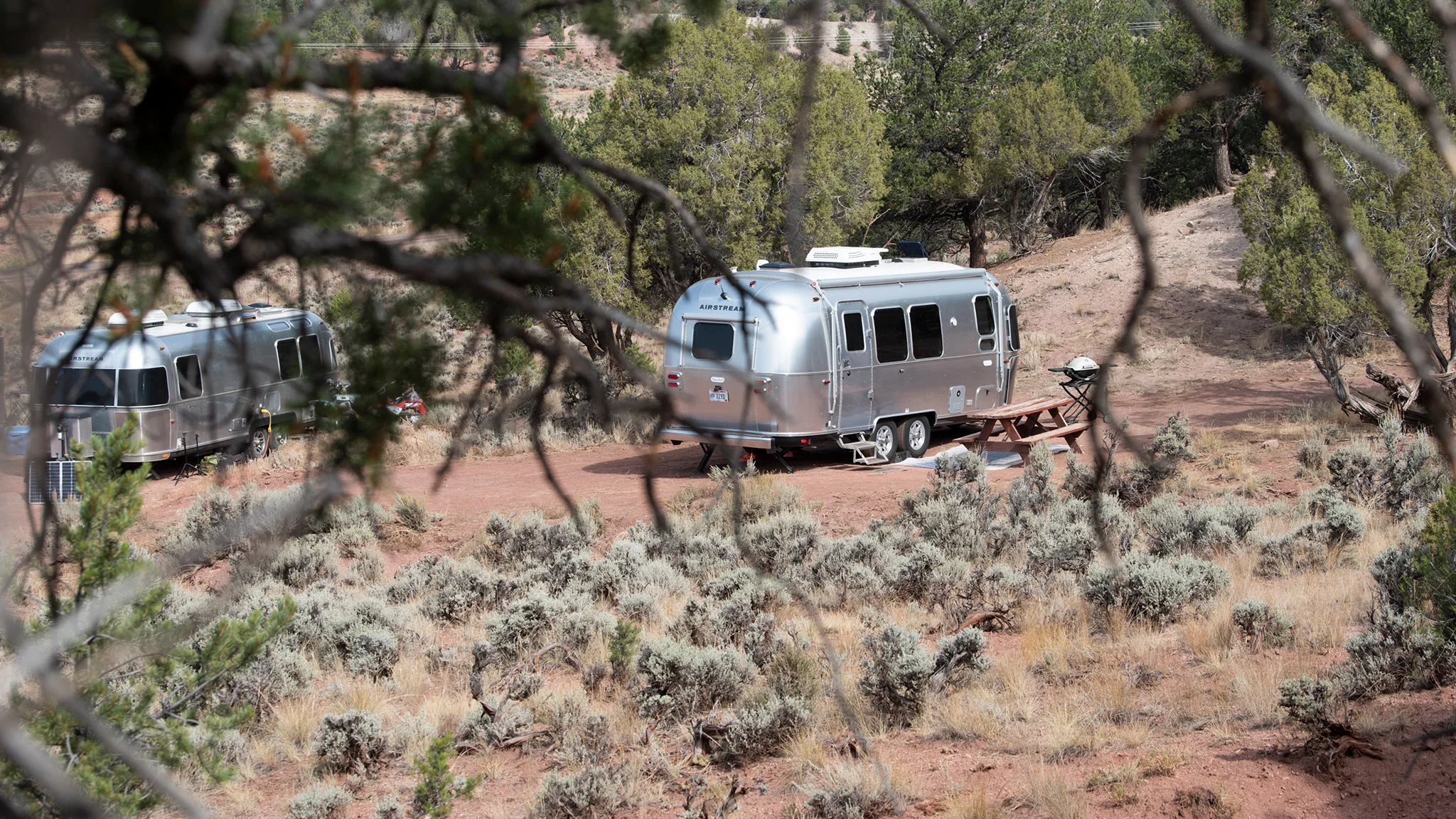 Two silver Airstream trailers are parked on a dirt clearing surrounded by dry brush, shrubs, and trees in a semi-arid landscape. A picnic table sits near one trailer, and branches frame the foreground.