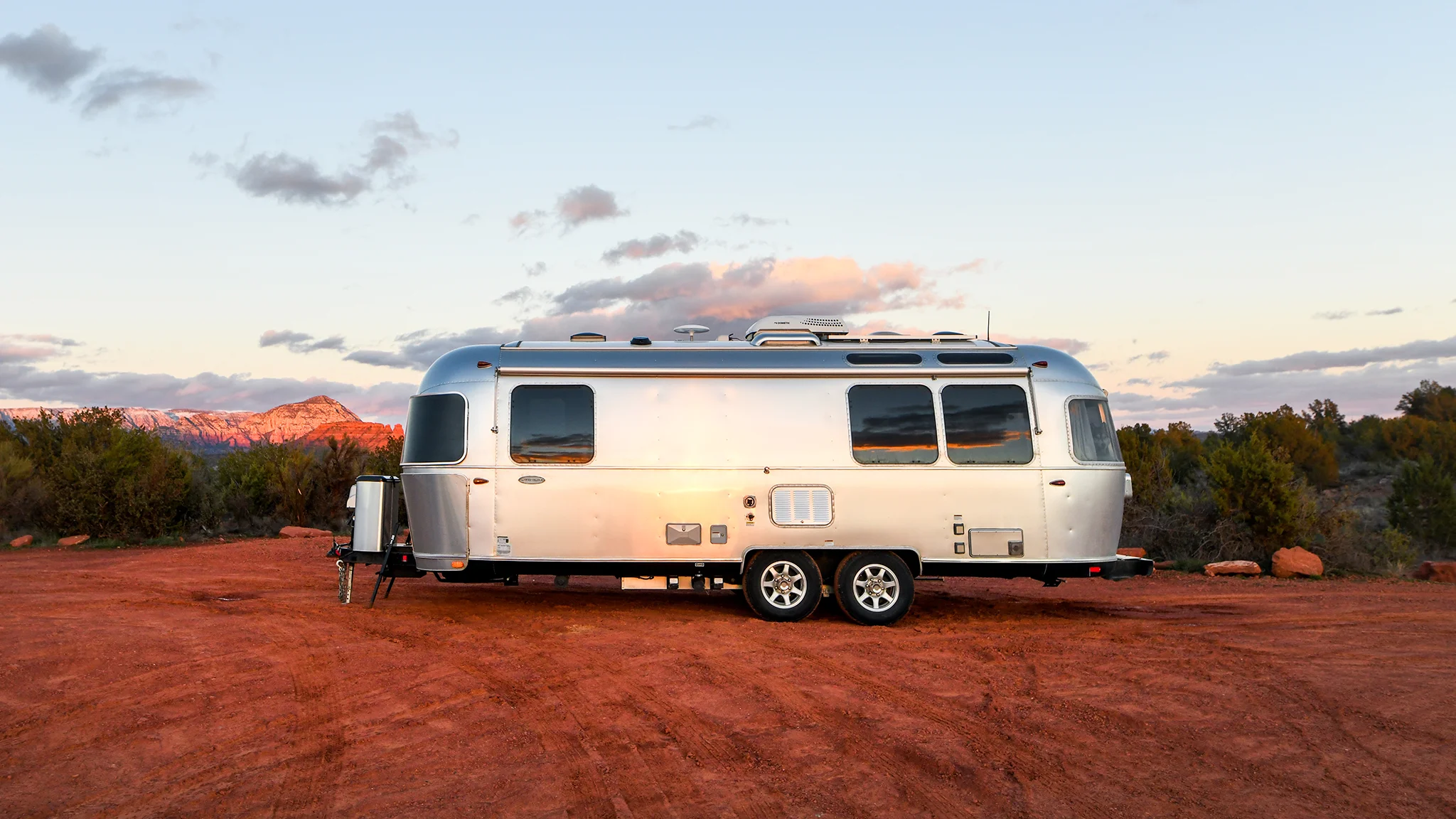 A shiny silver Airstream travel trailer is parked on red dirt, surrounded by desert shrubs, with mountains and a pastel sky in the background at sunset.