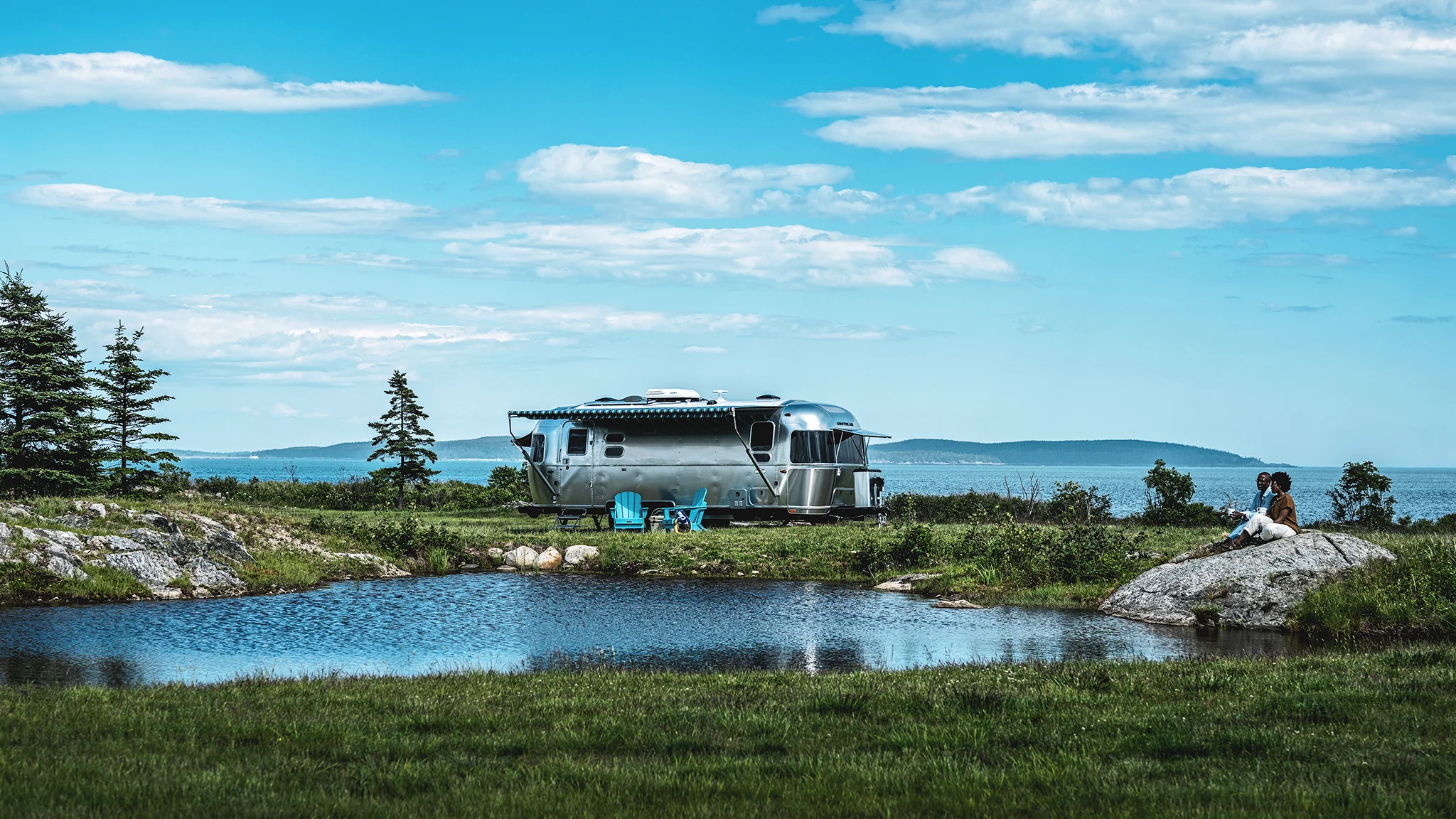 A silver RV parked near a small pond in a scenic landscape, with a person sitting on a rock and distant hills under a partly cloudy sky.