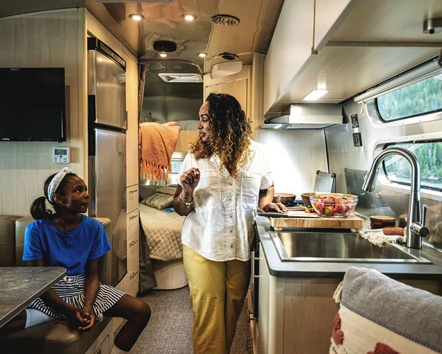 A woman and a girl sit and talk in a modern RV kitchen, featuring a table, sink, couch, and windows with scenic views outside.