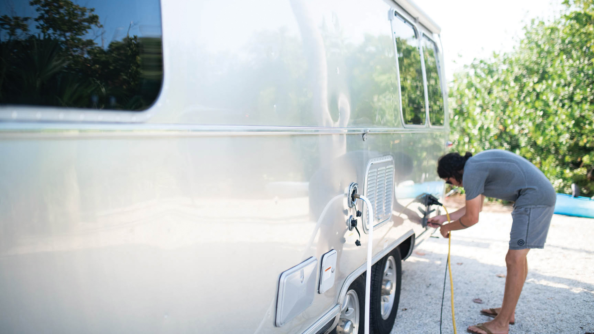 A person in shorts and a t-shirt washes the side of a shiny silver camper van with a yellow hose, surrounded by greenery and sunlight.