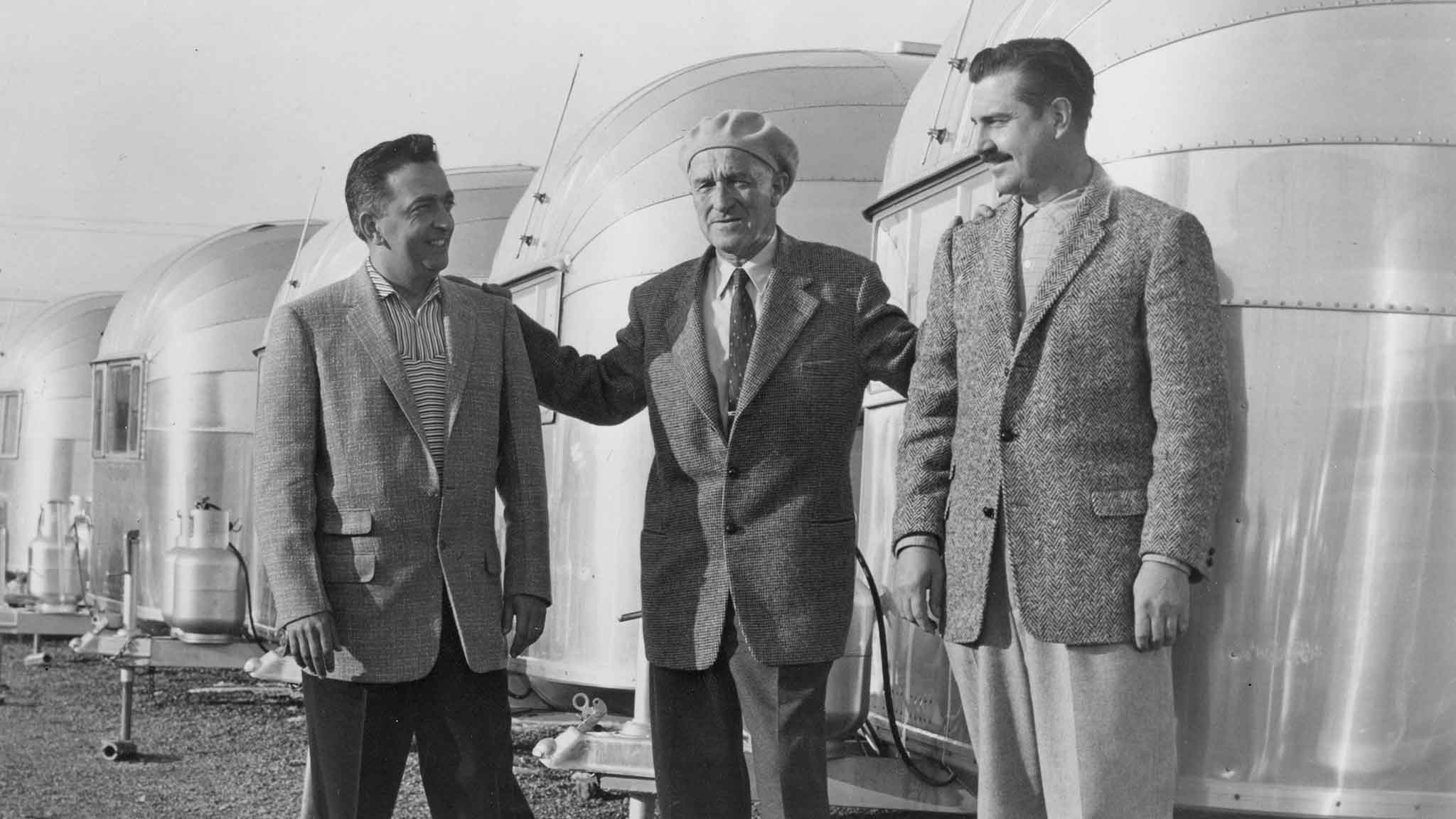 Three men in suits stand smiling in front of shiny, vintage trailers on a gravel surface.