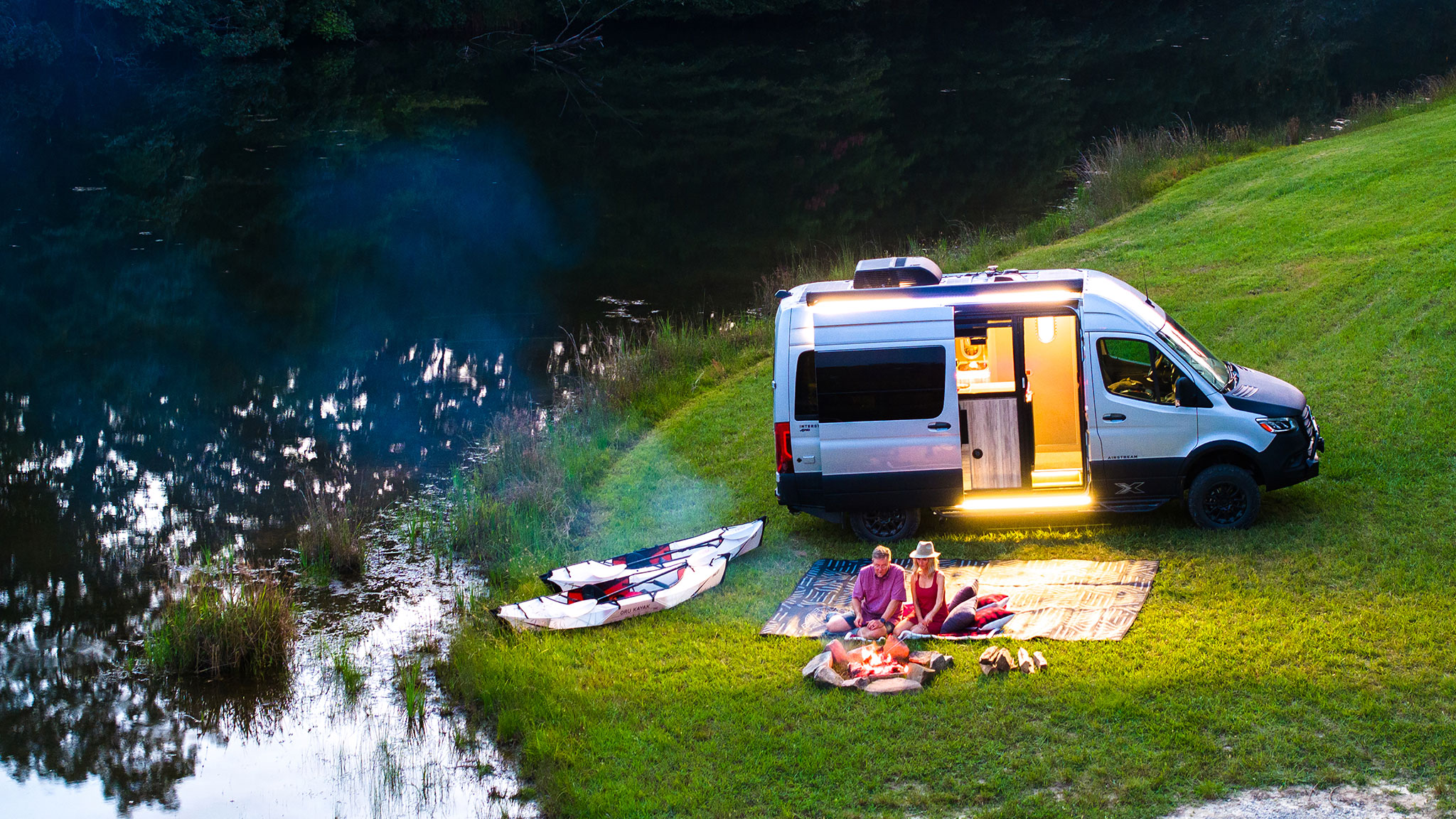 Camper van parked by a lake, with two people sitting on mats near a campfire and kayaks on the grass, during twilight.