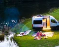 Camper van parked by a lake, with two people sitting on mats near a campfire and kayaks on the grass, during twilight.