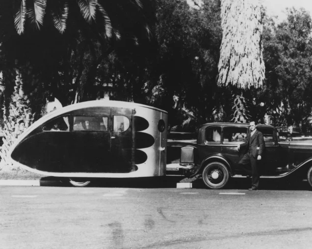 An Airstream Torpedo travel trailer being towed by a vintage car in a tropical area|An old Western Music and Radio Trades Journal|A vintage Airstream Torpedo travel trailer with people sitting outside of the trailer and car.|Two women sitting in lawn chairs next to an Airstream Torpedo travel trailer|An old clip from a newspaper about the Airstream Torpedo travel trailer|Dr. and Mrs. Holman holding up the Airstream Torpedo travel trailer blueprint while standing in front of the Airstream trailer|An Airstream Torpedo travel trailer being towed by a vintage car in a tropical area