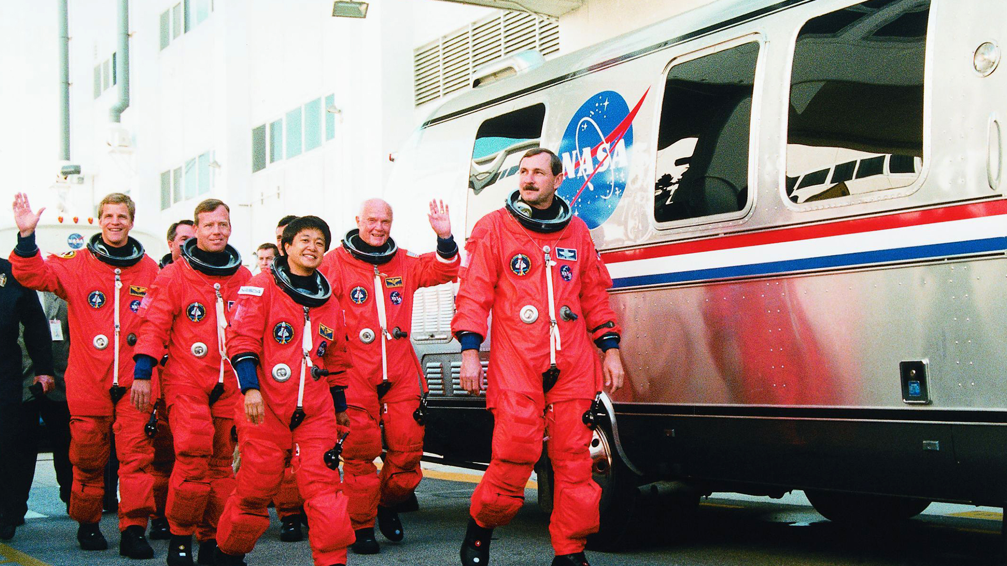 A group of astronauts in orange NASA spacesuits walk and wave beside a NASA vehicle, smiling as they head toward their space mission.