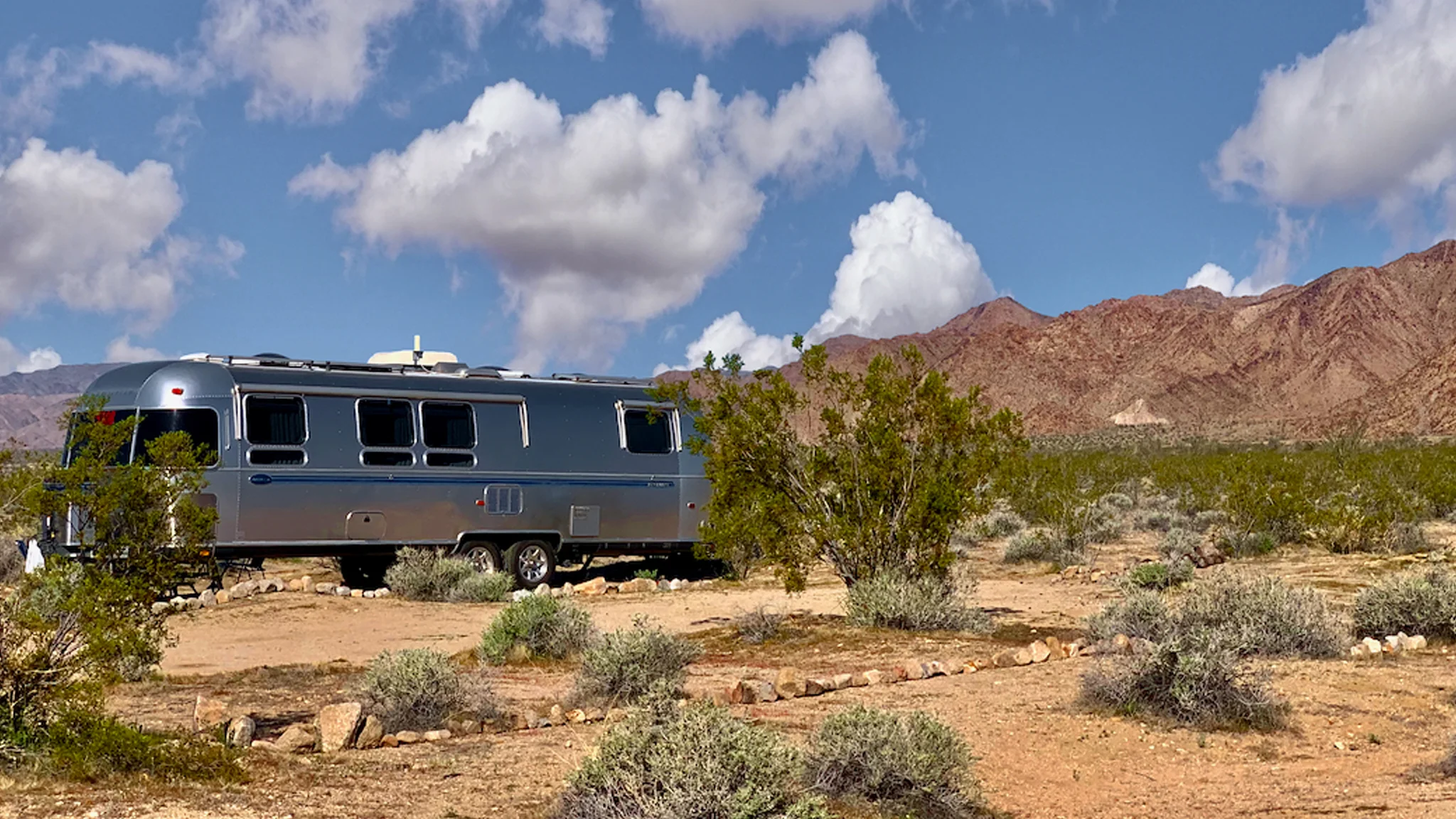 A silver Airstream trailer is parked in a desert landscape with scattered bushes, under a blue sky with fluffy clouds. Rugged, red mountains rise in the background.