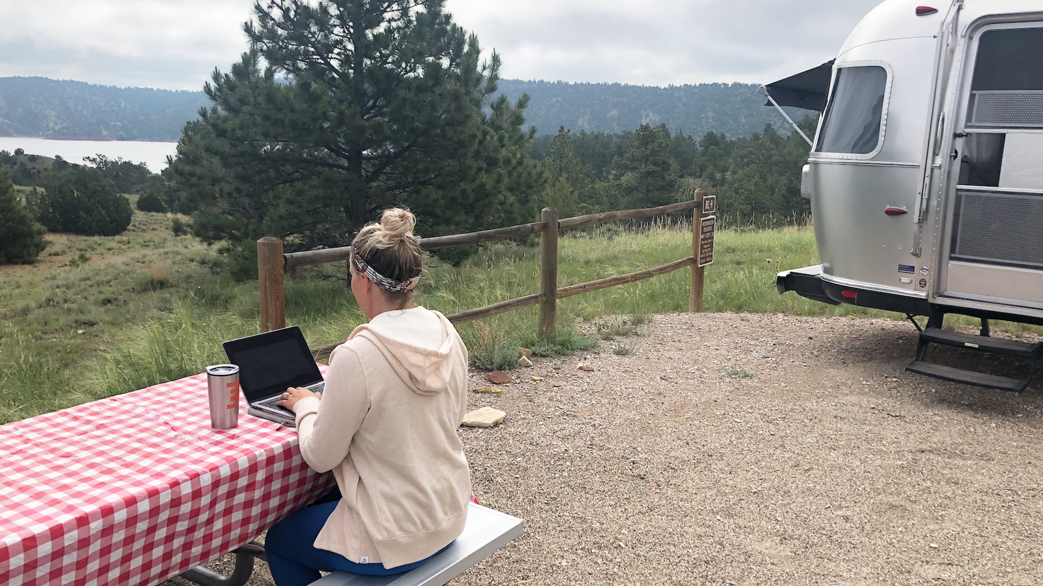 A person works on a laptop at a picnic table with a red checkered tablecloth near an Airstream trailer, overlooking a scenic, tree-filled landscape with hills and a lake in the background.