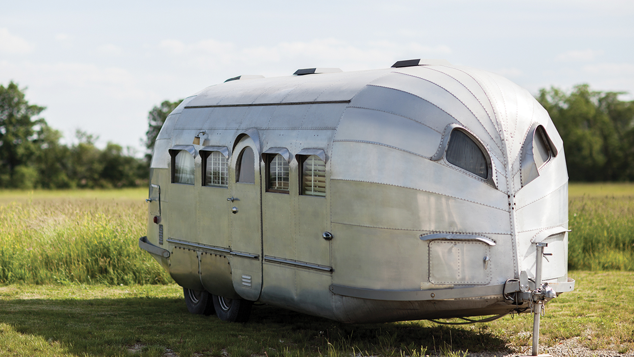 A vintage silver travel trailer with rounded edges and small windows sits on grassy land with trees and a blue sky in the background.