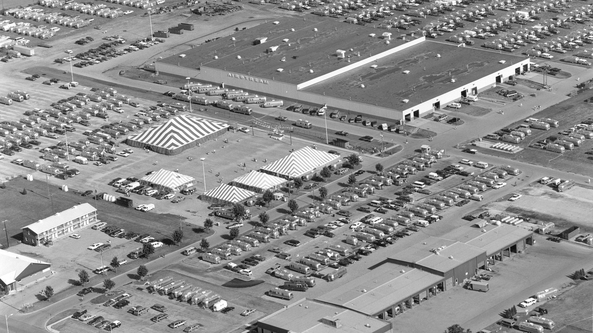 Aerial view of a large parking lot filled with cars, several striped tents, and a big commercial building. Smaller structures and more parked cars surround the main area. The image is in black and white.