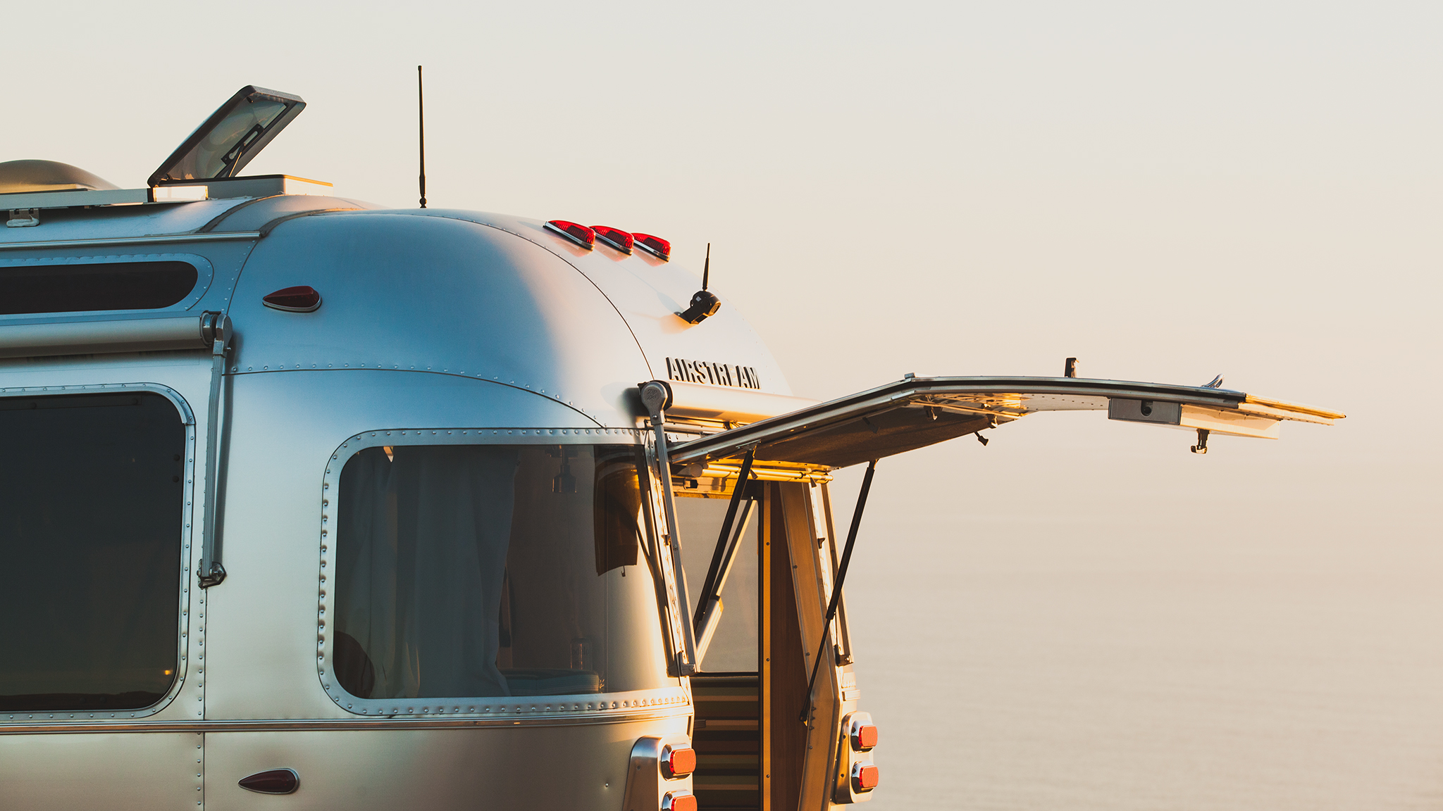 A shiny silver Airstream trailer with its rear hatch door open, parked near the ocean at sunset, with soft sunlight reflecting off its metallic surface.