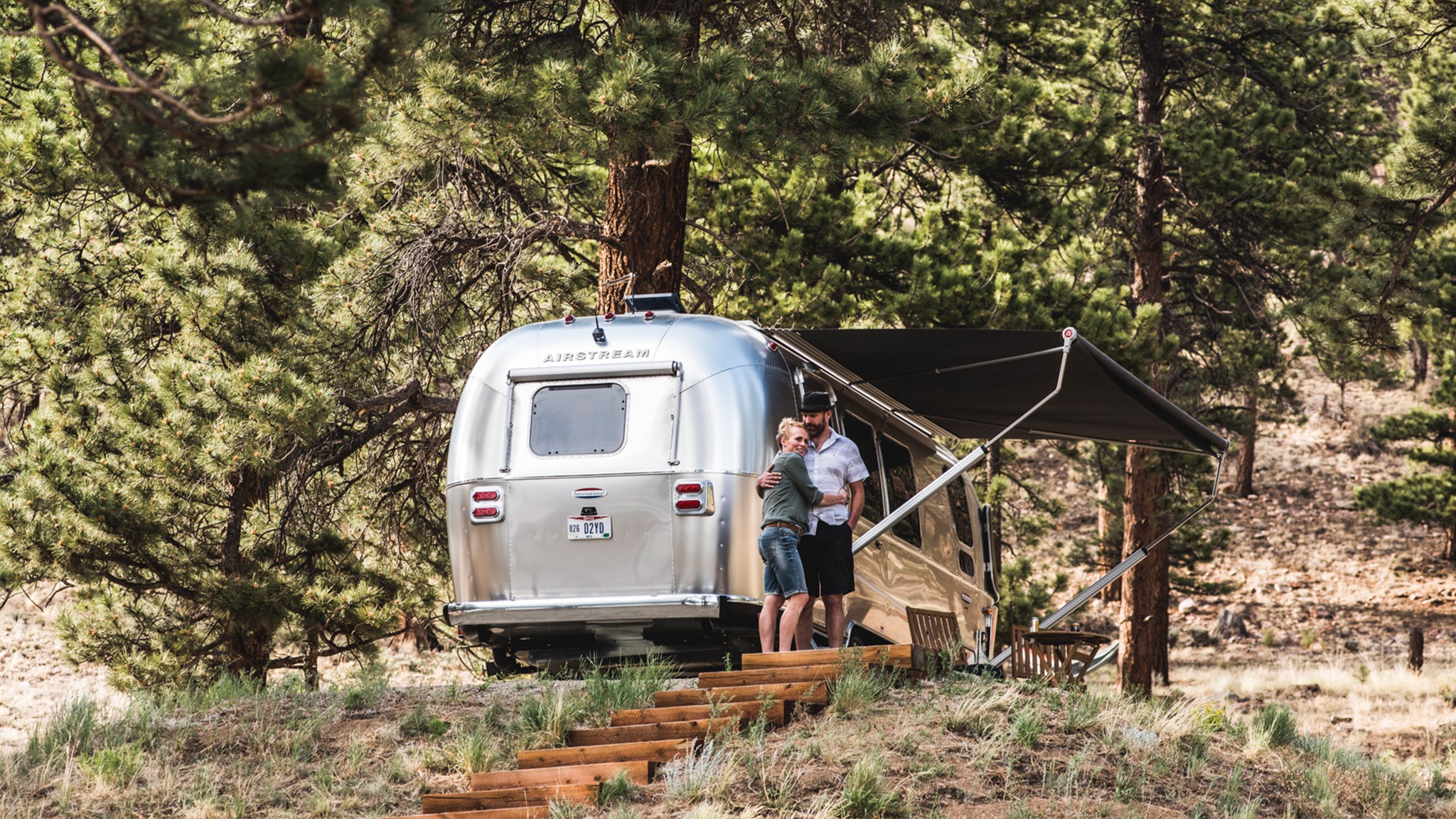 Two people stand hugging outside a silver Airstream trailer parked in a wooded area with tall pine trees. The trailer’s awning is extended, and wooden steps lead up to the campsite.