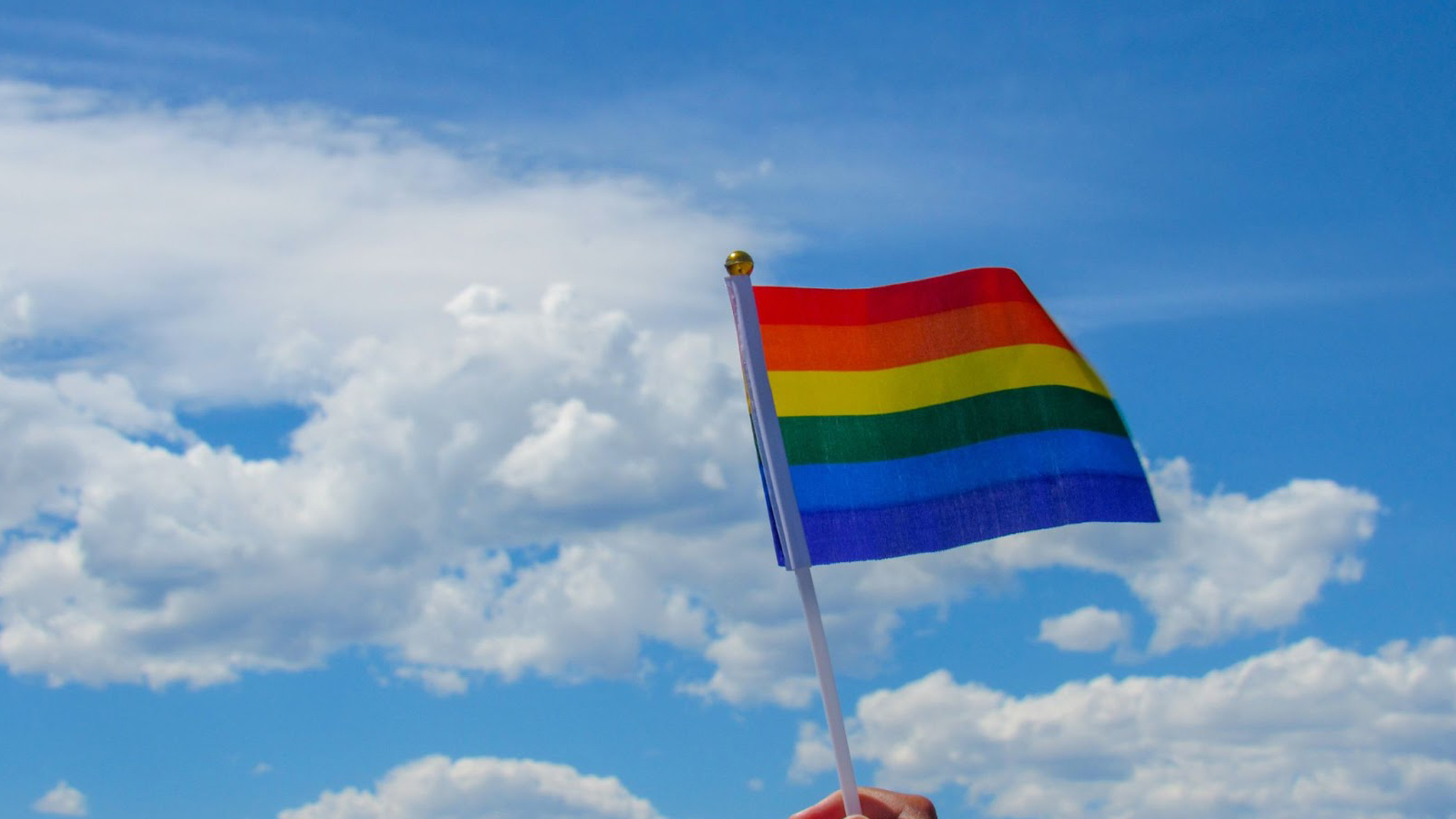A hand holds up a small rainbow Pride flag against a bright blue sky with scattered white clouds.
