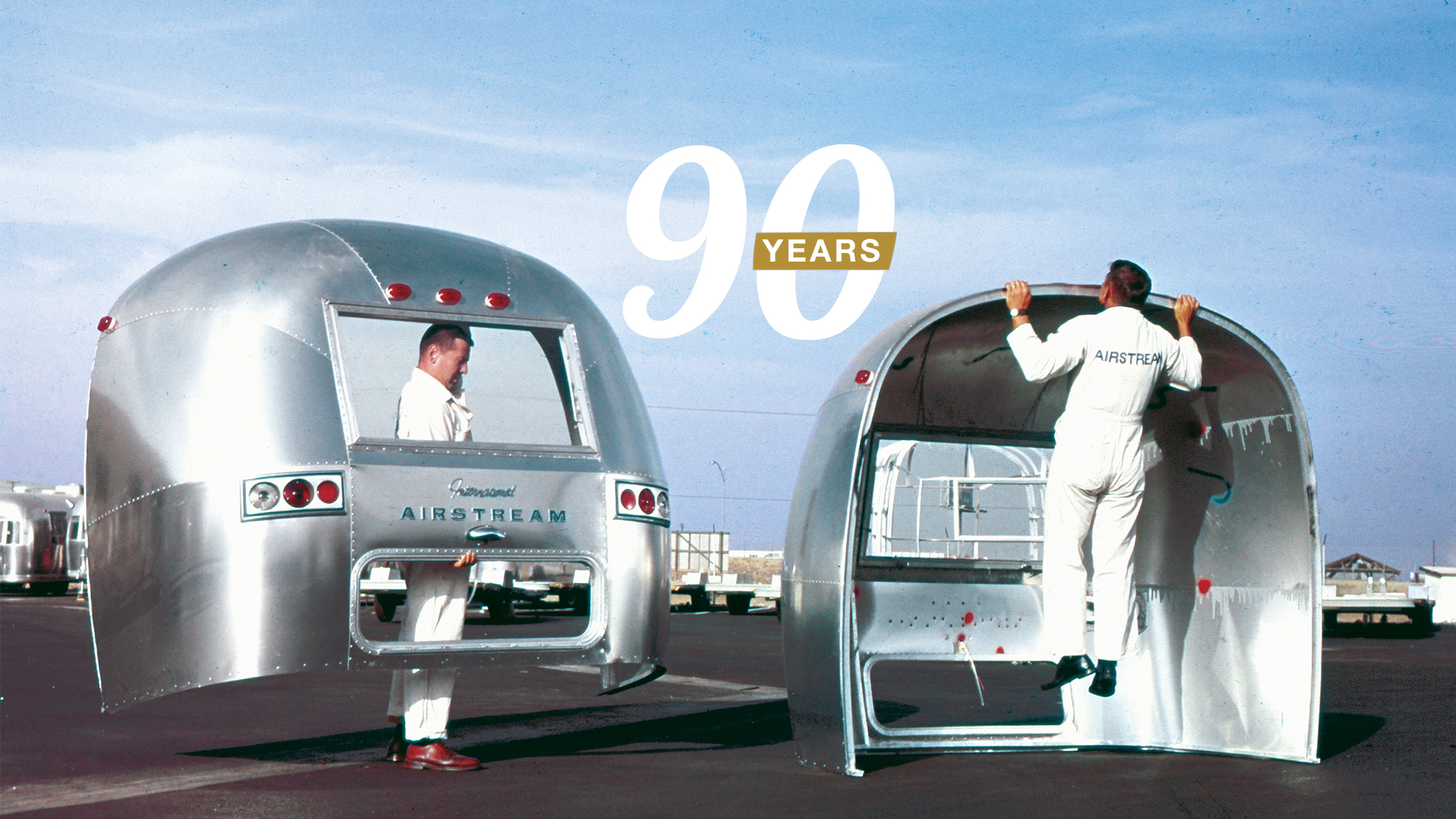 Two men in white coveralls stand inside lightweight, shell-like Airstream trailers, one lifting himself up, against a clear blue sky. Large white text reads “90 YEARS.”.
