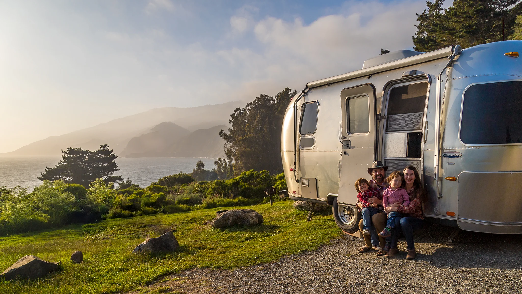 A family of three sits by their silver camper van parked on a scenic cliffside, with lush greenery, trees, and mountains overlooking the ocean at sunset.