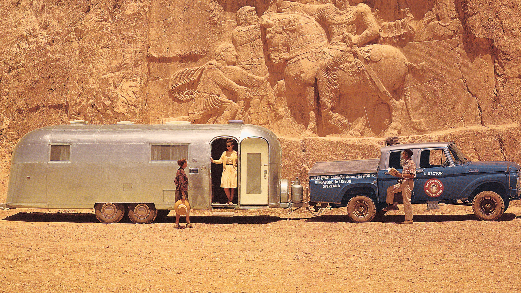 A silver trailer and blue truck are parked on sandy ground near an ancient rock carving. Three people stand around the vehicles. The scene appears sunny and warm, with a large stone relief in the background.