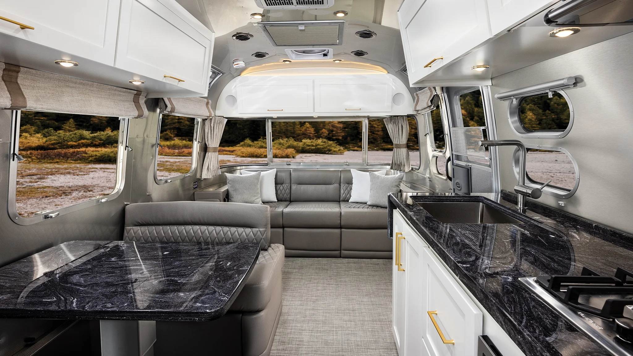 Modern RV interior with a marble-patterned kitchen, gray dining area, and large sofa. Large windows provide natural light and a view of outdoor trees. White cabinets and gold hardware create a sleek, stylish look.
