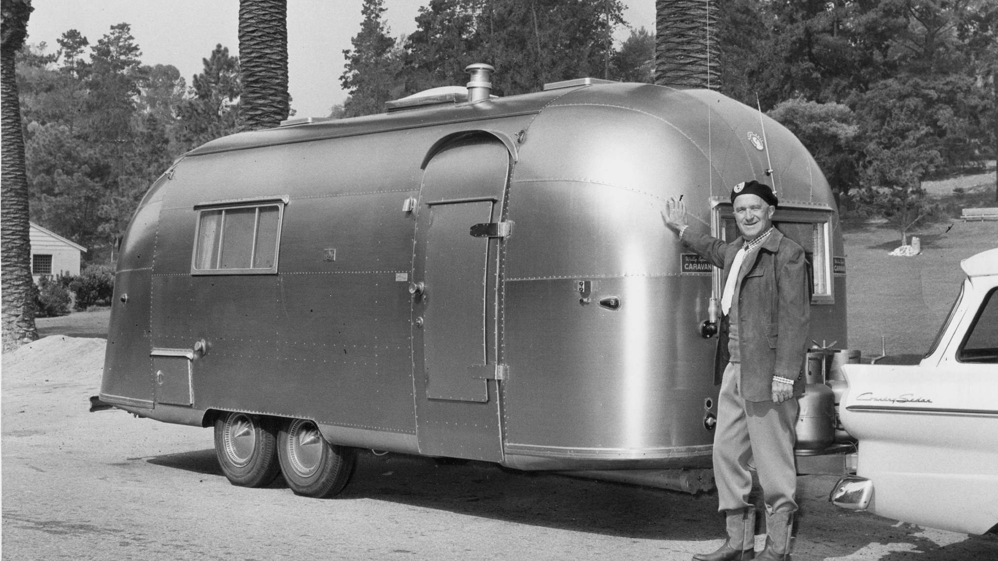 A man stands smiling and waving beside a polished, vintage Airstream trailer hitched to a classic car, with trees and a house visible in the background. The scene is in black and white.