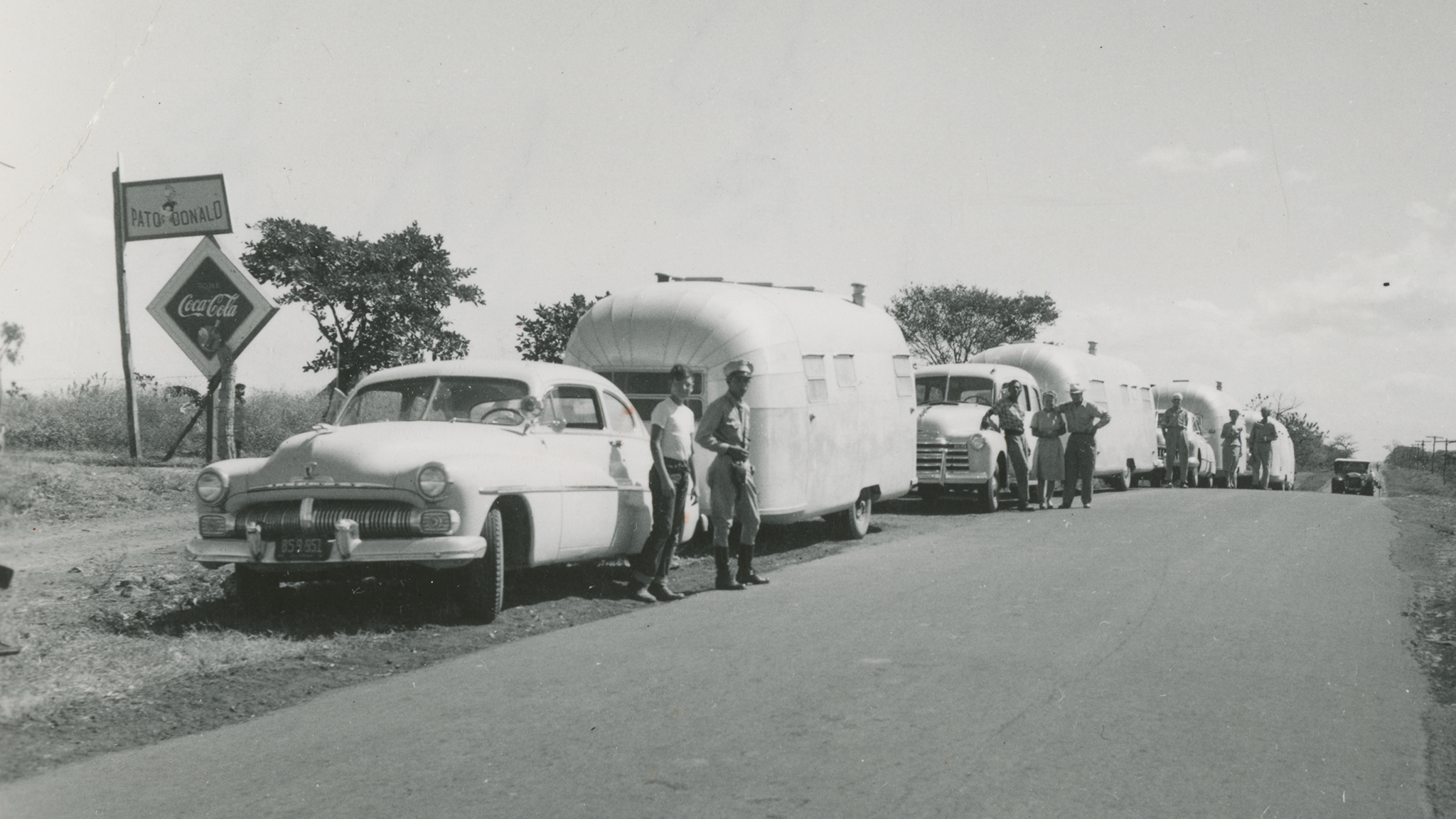 A black-and-white photo shows several vintage cars towing shiny travel trailers parked along a rural road. A few people stand beside the vehicles. A roadside sign reads “Auto Coronado” with a Coca-Cola logo. Trees and open sky are visible.