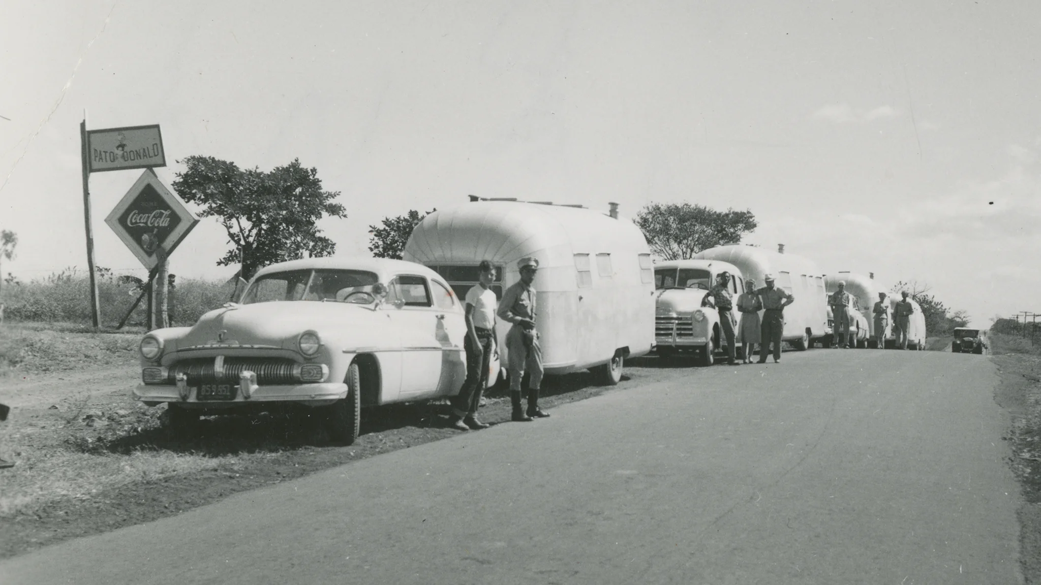 A black-and-white photo shows several vintage cars towing shiny travel trailers parked along a rural road. A few people stand beside the vehicles. A roadside sign reads “Auto Coronado” with a Coca-Cola logo. Trees and open sky are visible.