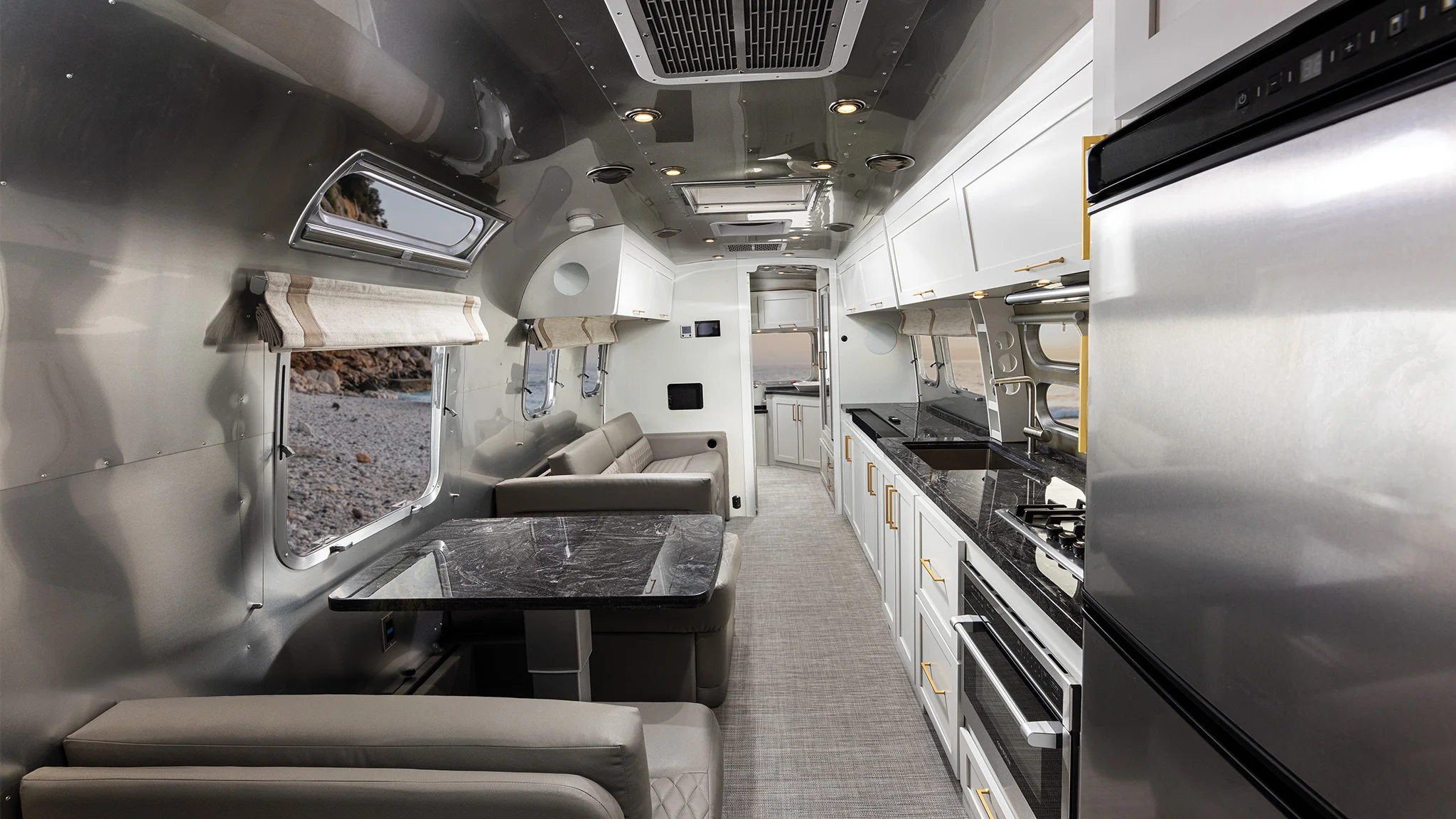 Interior of a modern RV with sleek metallic walls, a kitchenette, sofa, dining area, and large windows showing an outdoor scene.