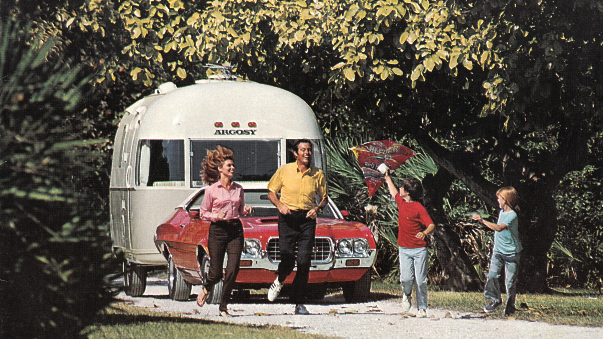 A family of four joyfully runs beside a red car towing an Argosy travel trailer on a sunny day, surrounded by lush greenery; one child holds a colorful kite.