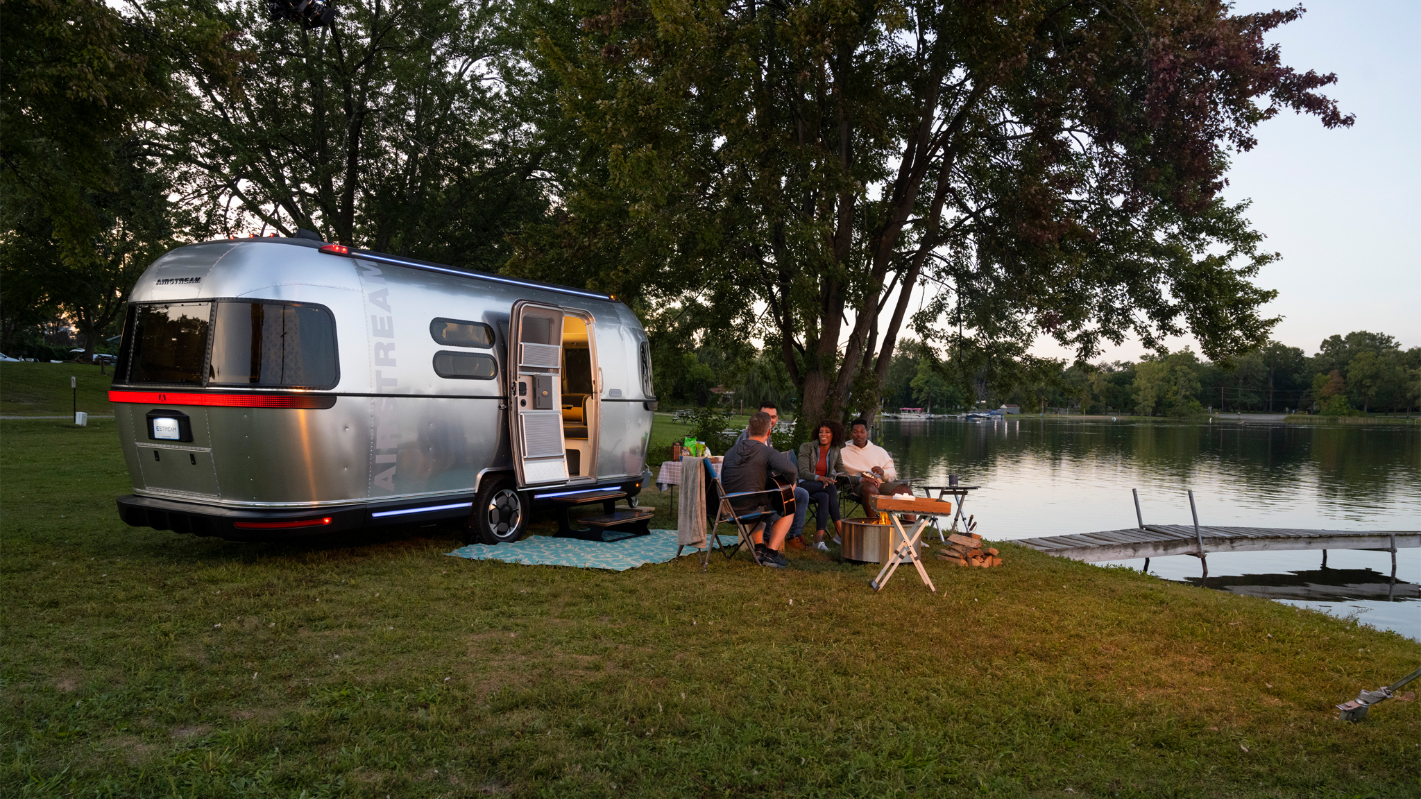 A group of people sit around a table near a lake, next to a silver Airstream trailer under trees. The sun is setting, and a wooden dock extends into the calm water in the background.