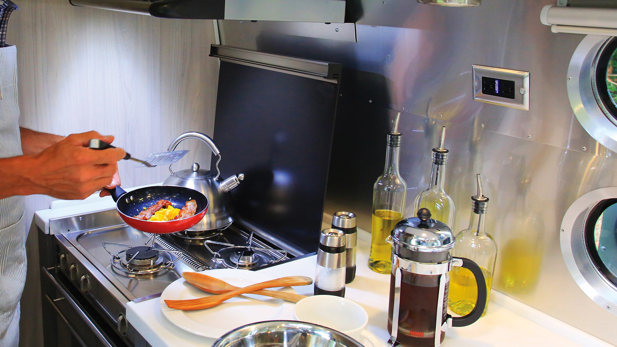 A person cooks in a modern kitchen, holding a frying pan with food over a stove. Nearby are kitchen utensils, oil bottles, a kettle, a French press with coffee, and two round windows on the metallic wall.