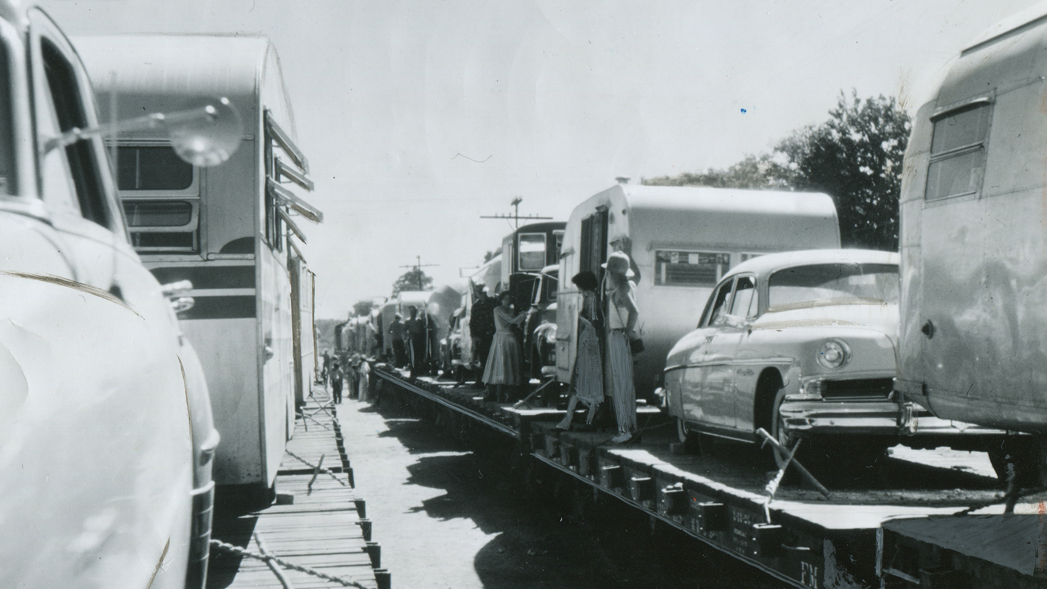 Black-and-white photo of vintage cars and trailers loaded on a train flatbed, with people standing between the vehicles and the train car, and trees visible in the background.