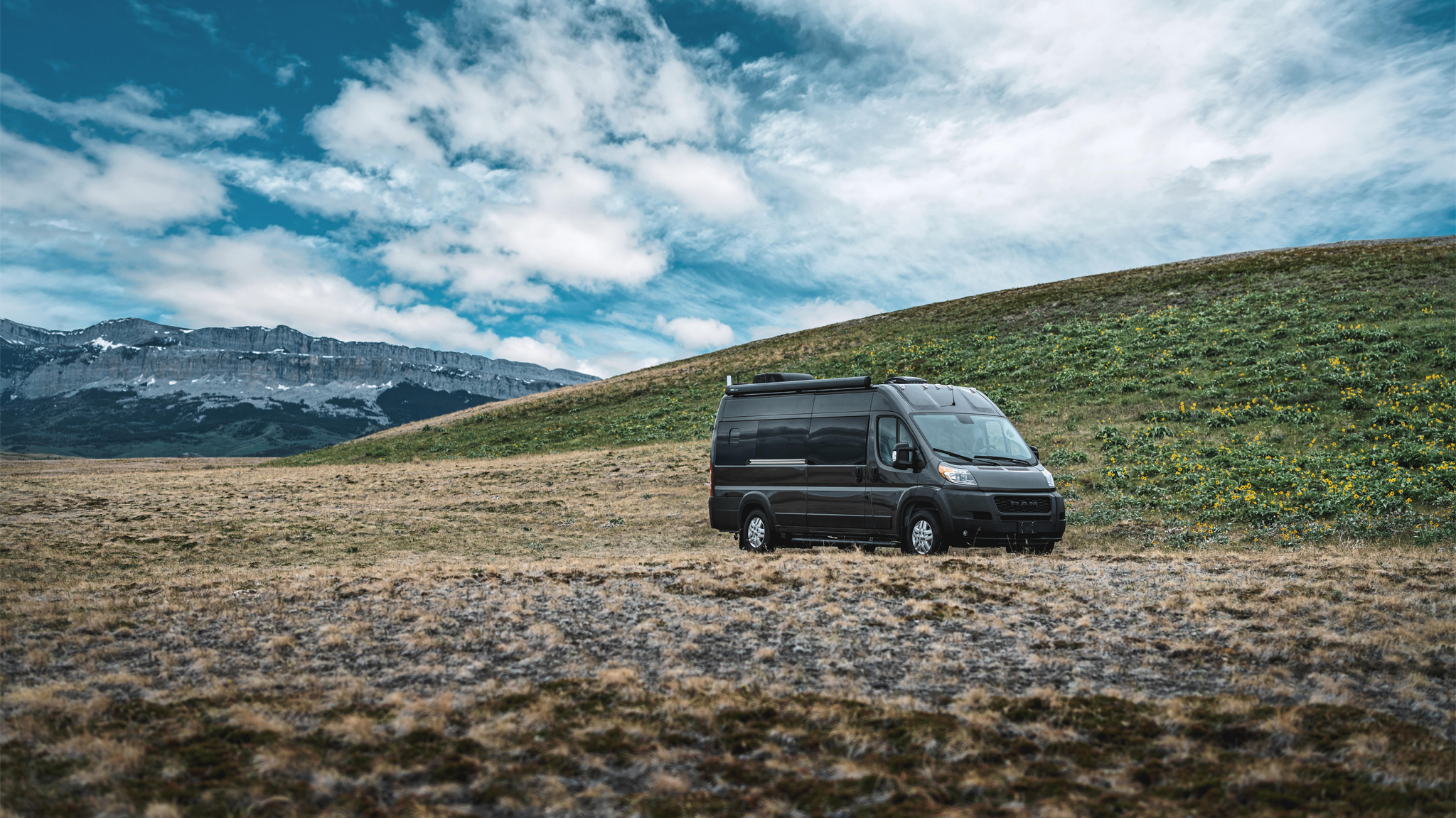 A black camper van is parked on a grassy, slightly sloped hillside with mountains in the background under a partly cloudy sky. Yellow flowers dot the green hill.