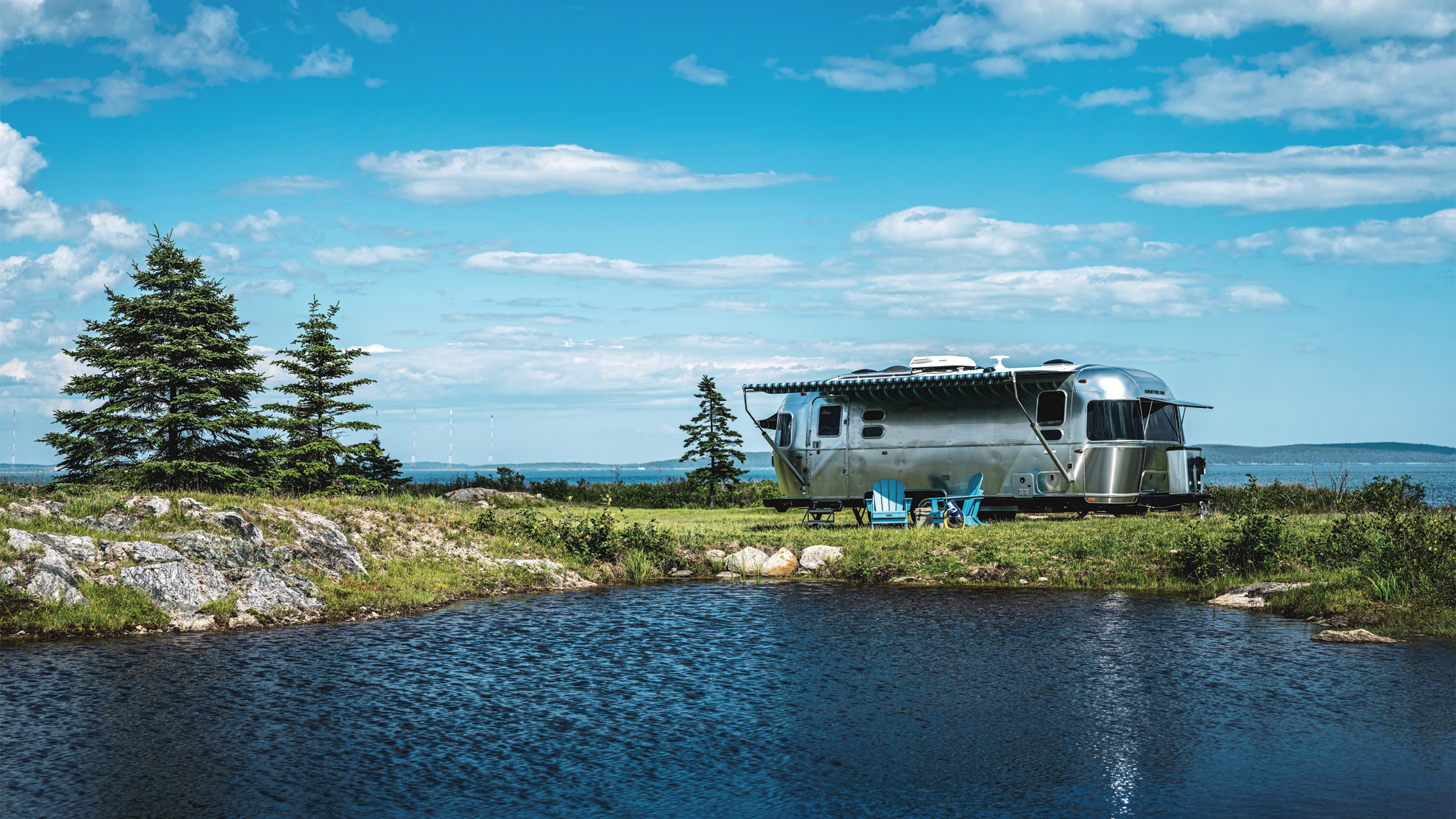 A silver camper trailer is parked by a pond with blue water, surrounded by grass, rocks, and a few pine trees under a partly cloudy blue sky. Blue chairs are set up next to the trailer.