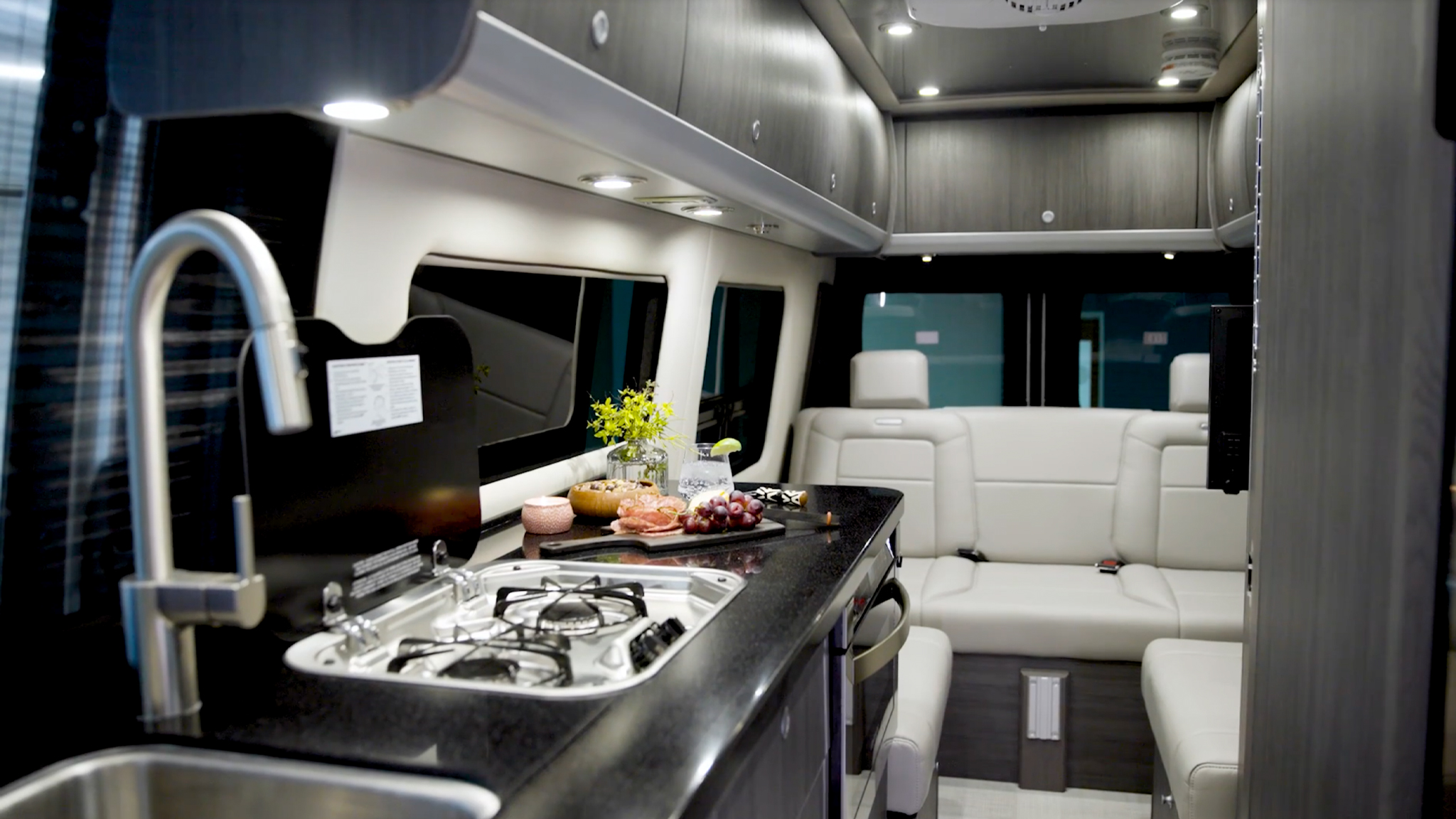 Luxurious RV interior featuring a sleek kitchen with a stove and sink, black counter, and a comfortable white seating area with soft lighting.