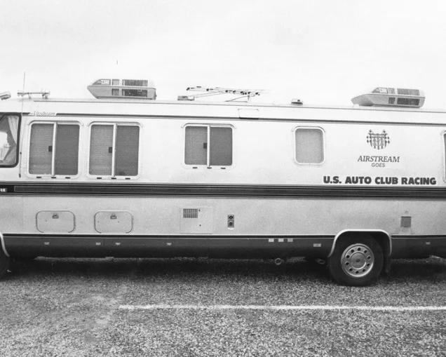 A vintage Airstream motorhome with "U.S. Auto Club Racing" lettering, parked on gravel, featuring multiple windows and rooftop units.