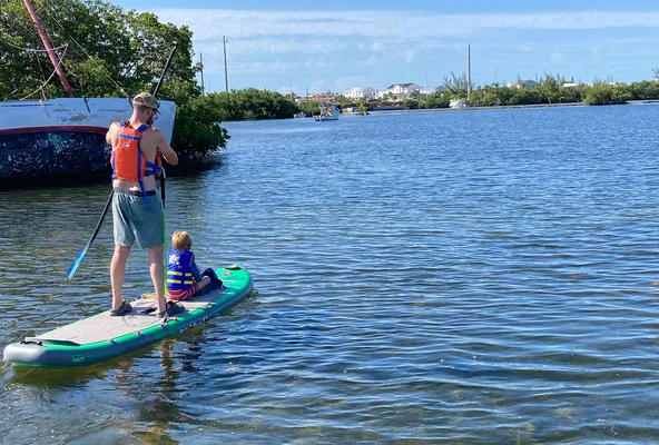 A man and child paddleboarding on a calm lake, surrounded by trees and distant buildings under a clear blue sky.