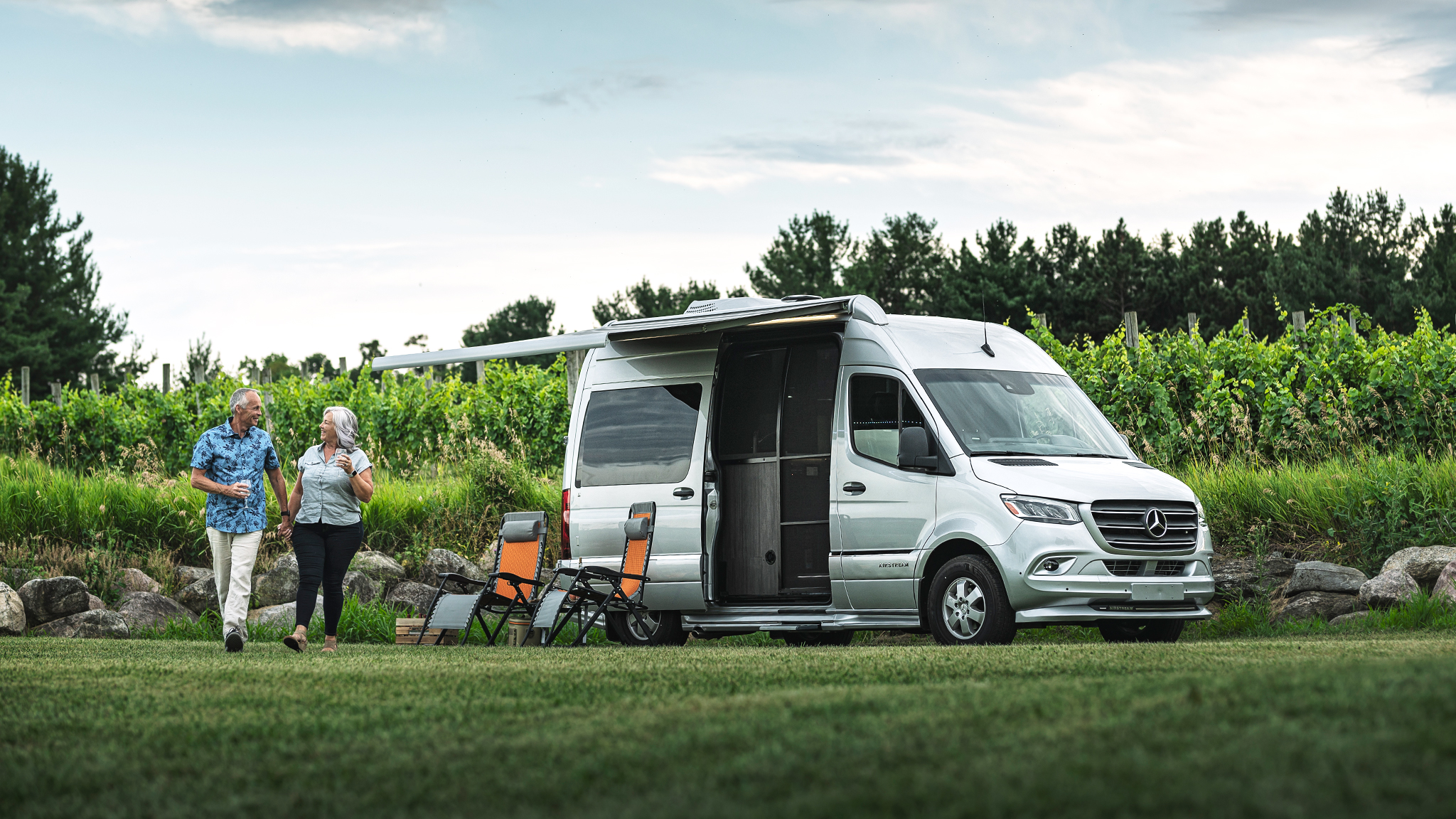 Elderly couple walking hand in hand near a silver camper van, with foldable chairs and lush greenery in the background.