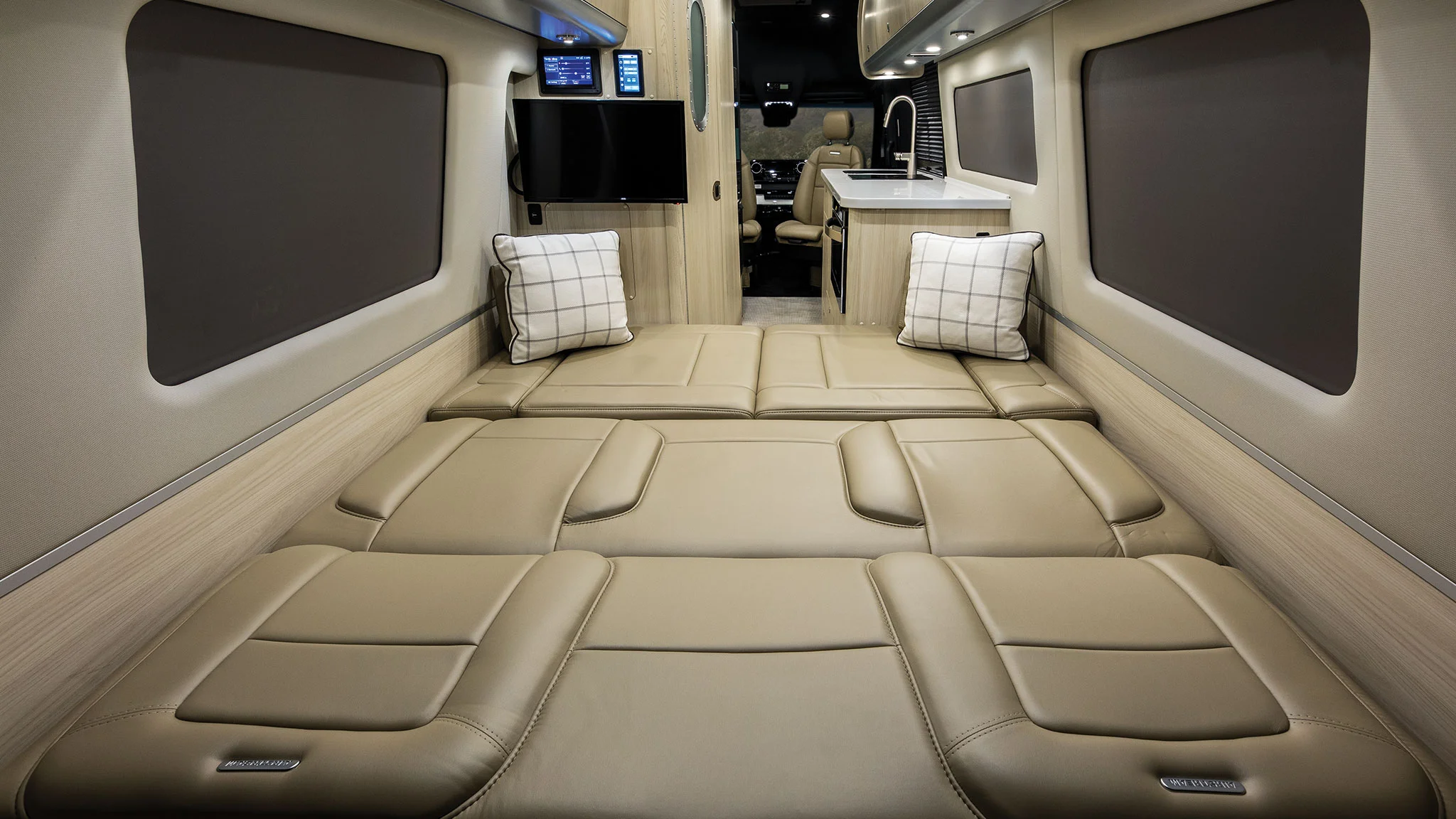 Luxurious RV interior with a spacious beige leather bed setup, two plaid pillows, wall-mounted screens, and ambient lighting.