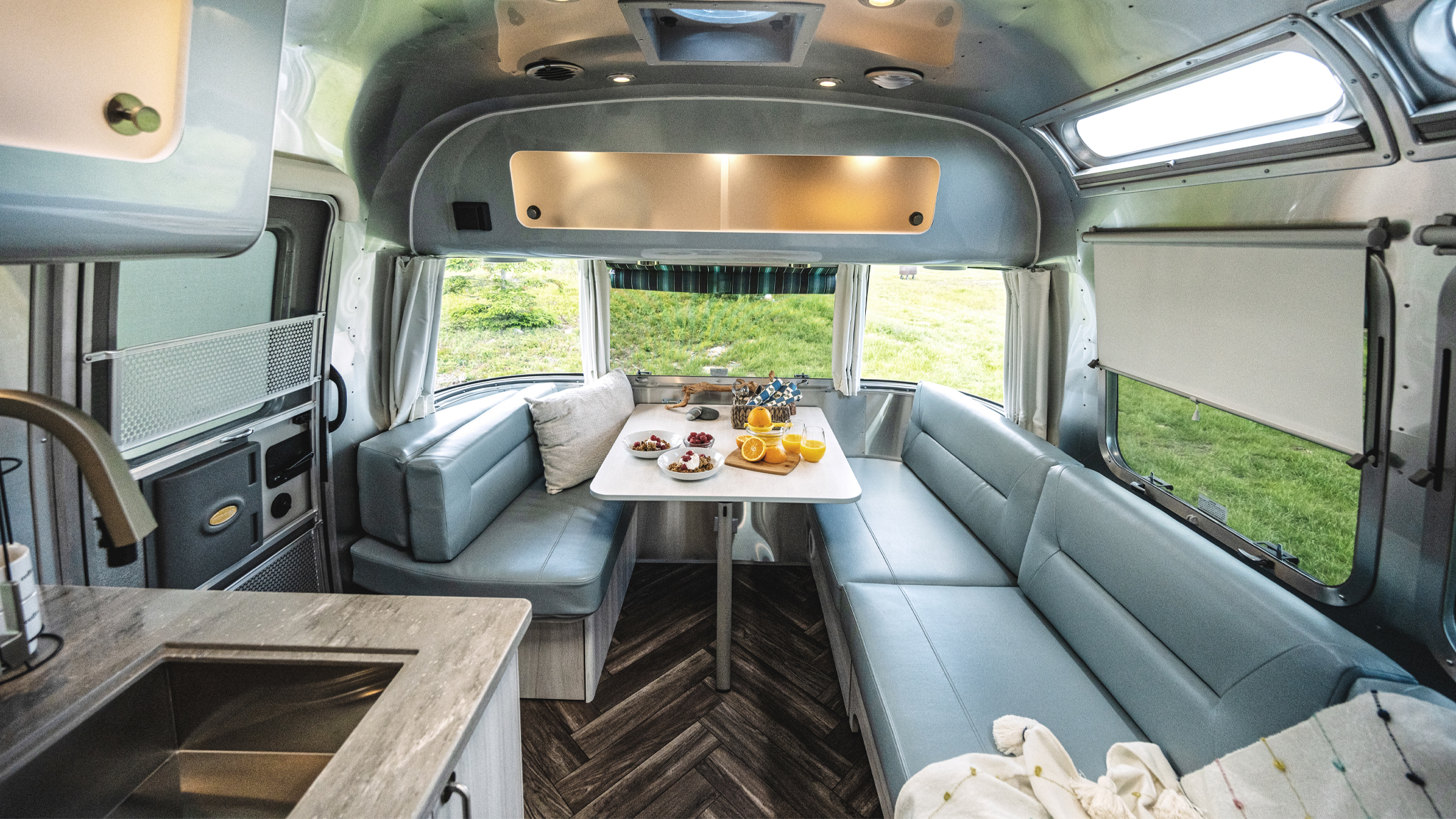 Interior of a modern camper van with light blue seating, a table set with breakfast items like juice and fruit, large windows showing green grass outside, and a compact kitchen area with a sink.